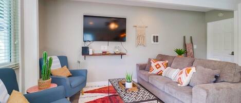 A living room with a grey sofa, a blue armchair, a coffee table, and a flat-screen TV mounted on the wall.