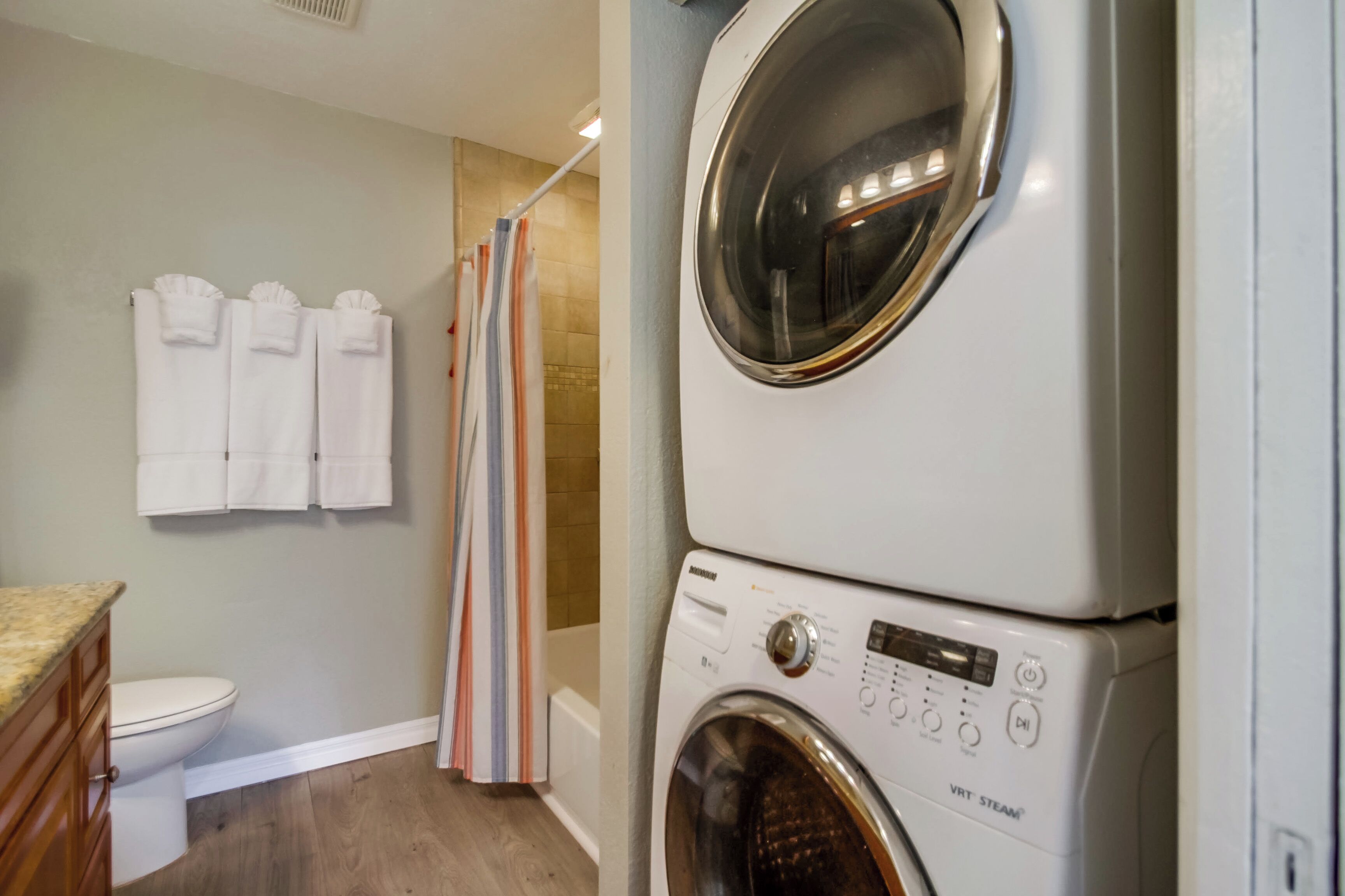 A laundry area with a front-loading washer and dryer, a shower with a striped curtain, and a toilet.