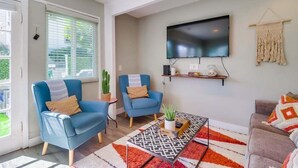 A living room with two blue armchairs, a coffee table with a geometric rug, a flat-screen TV mounted on the wall, and a macrame wall hanging. - Casa Azul Lower (San Diego)