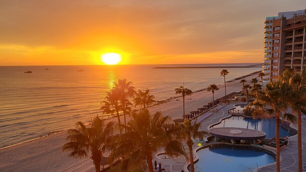 A resort with a pool, palm trees, and a view of the sunset over the ocean. - Southern Comfort at the Sonoran Sun (Puerto Peñasco)