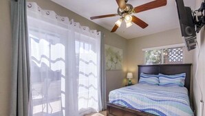 Une chambre à coucher avec un lit, un ventilateur de plafond, une fenêtre avec des stores et une chaise près de la fenêtre.
