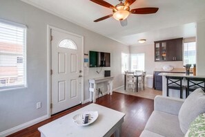 Sala de estar con sofá blanco, una mesa de centro y un comedor con mesa y sillas. Se observa una cocina con armarios y encimera.