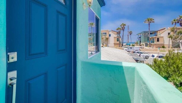 Une porte bleue avec une fenêtre et un luminaire, donnant sur une rue bordée de voitures garées et de palmiers.