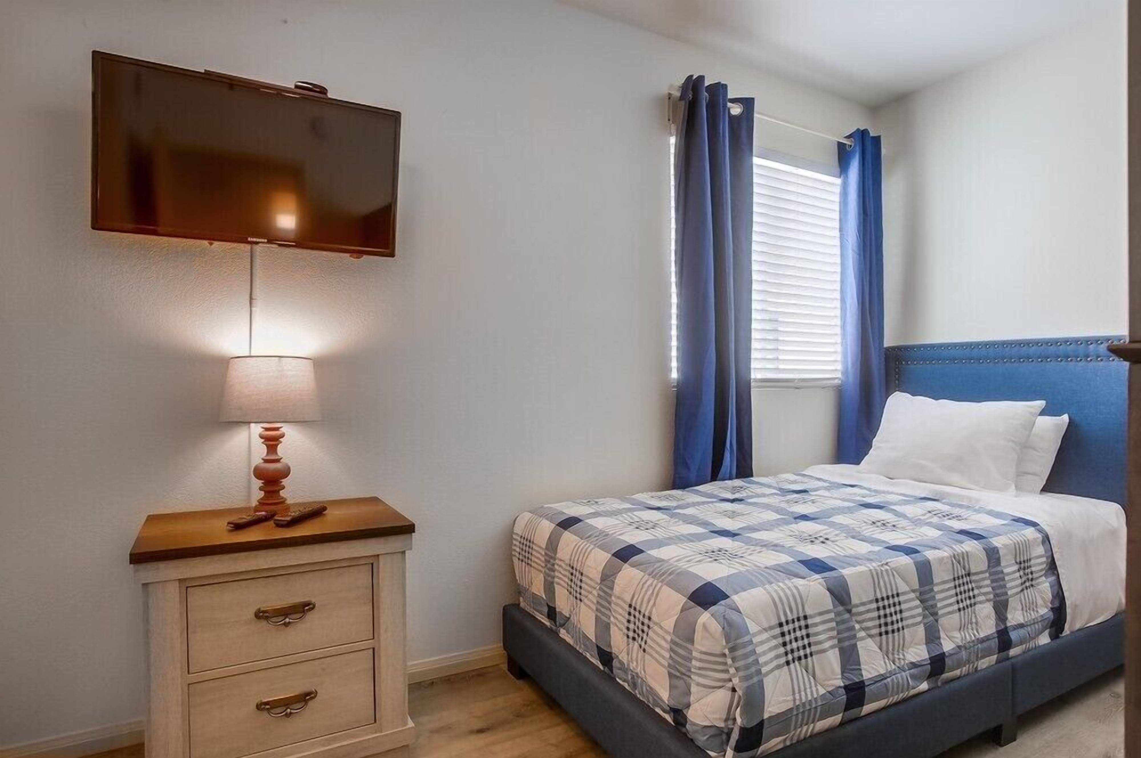 Une chambre à coucher avec un lit, un téléviseur fixé au mur, une table de chevet avec une lampe et une fenêtre avec des rideaux.