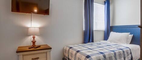 Une chambre à coucher avec un lit, un téléviseur fixé au mur, une table de chevet avec une lampe et une fenêtre avec des rideaux.