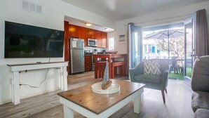 Una sala de estar moderna con un televisor de pantalla plana, una mesa de centro de madera y un sillón azul. Se ve una cocina con armarios de madera y un comedor con una mesa y sillas a través de una puerta abierta.