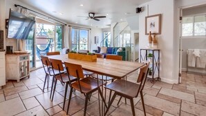 A spacious dining area with a wooden table and chairs, a ceiling fan, and large windows offering a view of the outdoors. - Ocean Breeze 1 (San Diego)