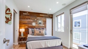 A bedroom with a wooden accent wall, a bed with pillows, a nightstand with a lamp, and a view of the outside. - Ocean Breeze 1 (San Diego)