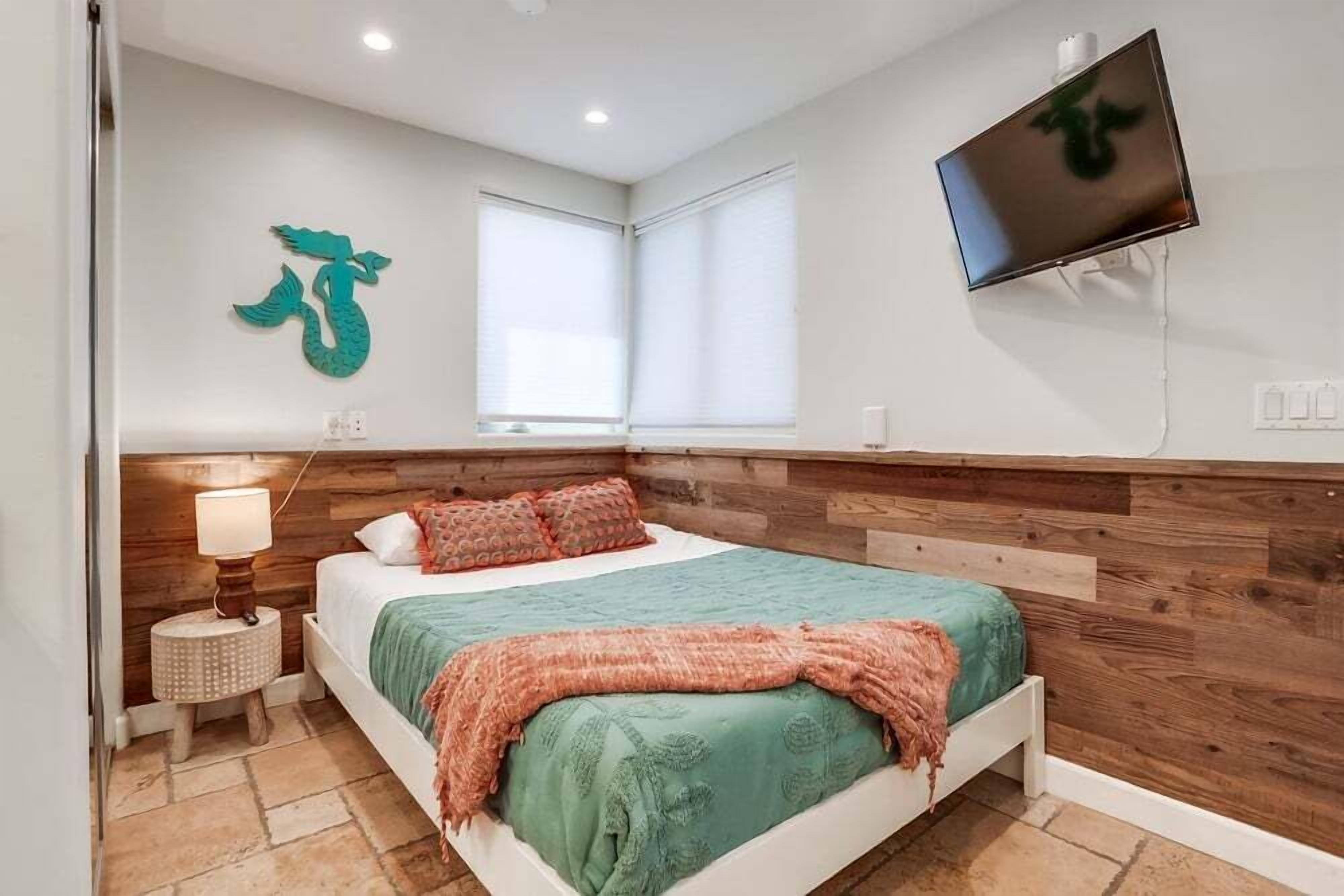 Un dormitorio con cabecera de madera, cama con colcha verde, decoración de sirena en color verde azulado, televisor montado en la pared y ventana con persianas.