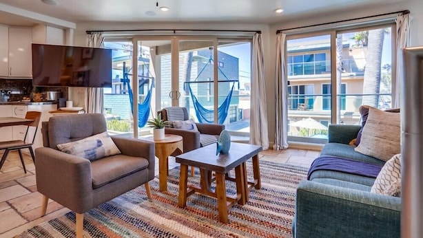 A modern living room with a sofa, armchair, and coffee table. There is a television mounted on the wall and a ceiling fan. Large windows provide a view of the outside. - Ocean Breeze 1 (San Diego)