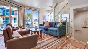 A modern living room with a sofa, armchairs, and a coffee table. There is a large window with curtains, a ceiling fan, and a view of buildings outside. - Ocean Breeze 1 (San Diego)