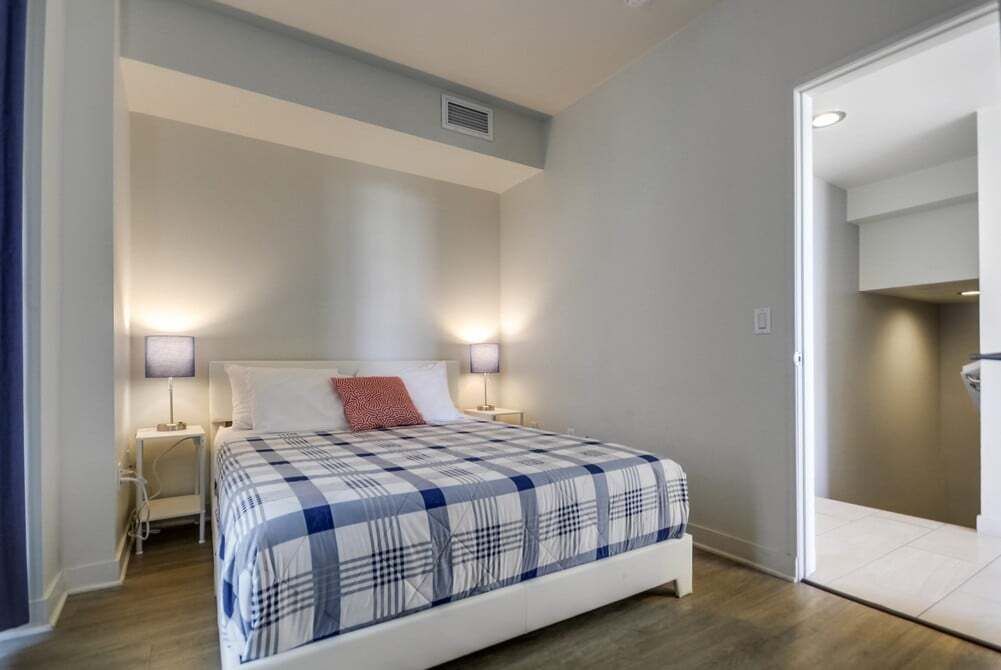 A bedroom with a bed, two bedside lamps, a nightstand, and a door leading to another room.