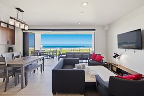 A modern living room with a dining area, a sofa set, a TV mounted on the wall, and a large window offering a view of the ocean. - Ocean Park 02 (San Diego)