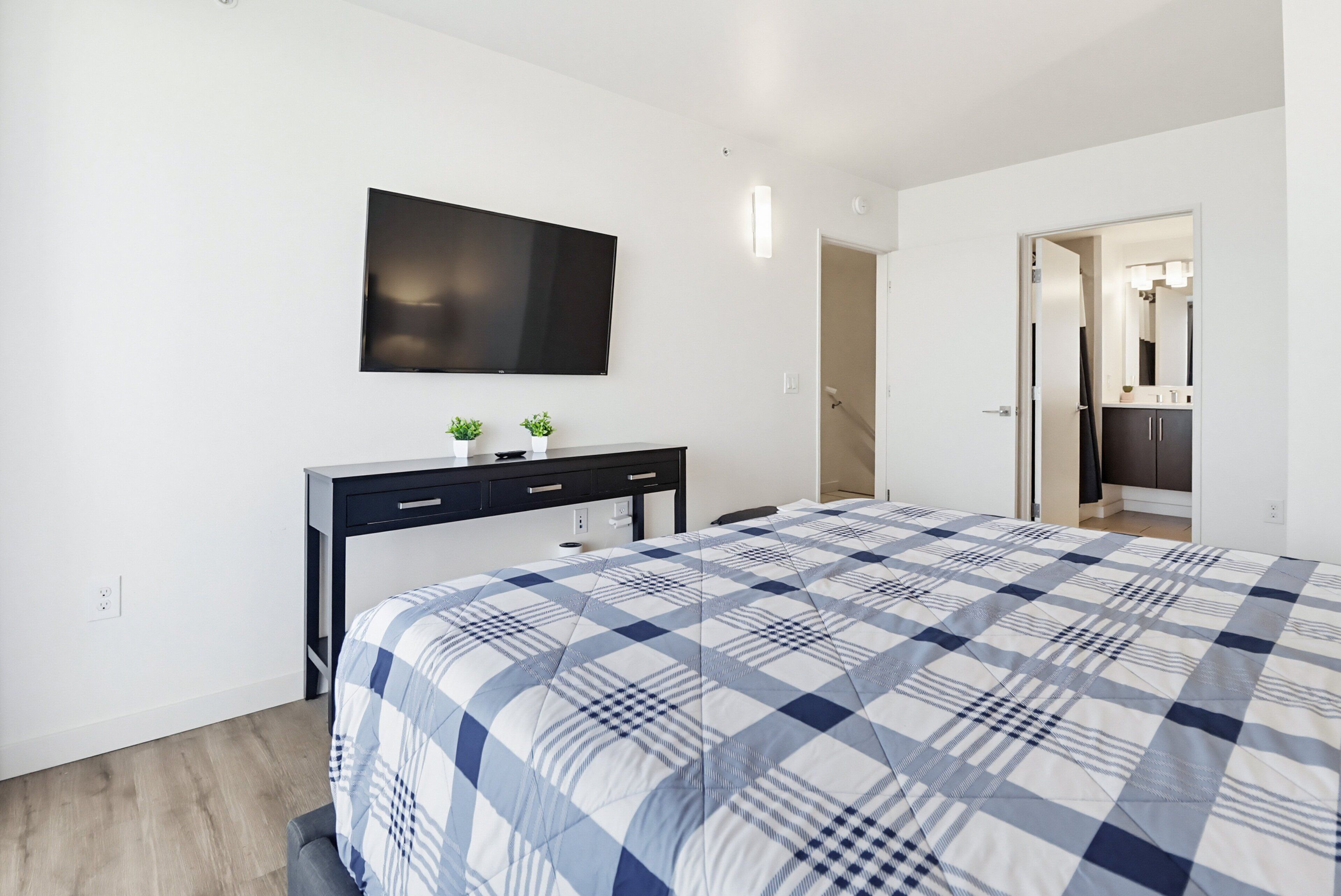 Une chambre à coucher comprenant un lit, un téléviseur fixé au mur, un bureau avec des tiroirs et une porte donnant sur une autre pièce.