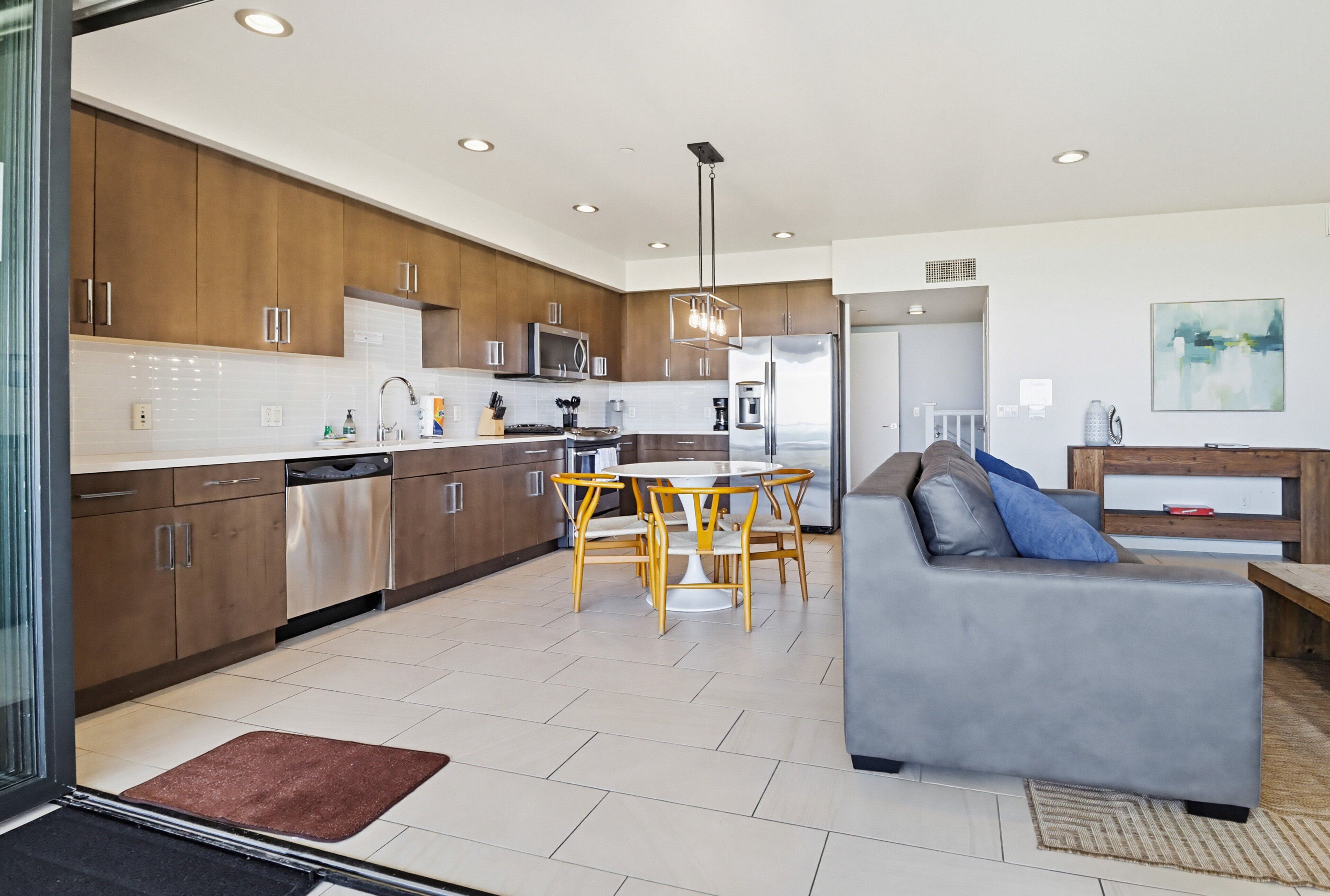 A modern kitchen with wooden cabinets, a dining area with a round table and chairs, and a grey sofa in a living room.
