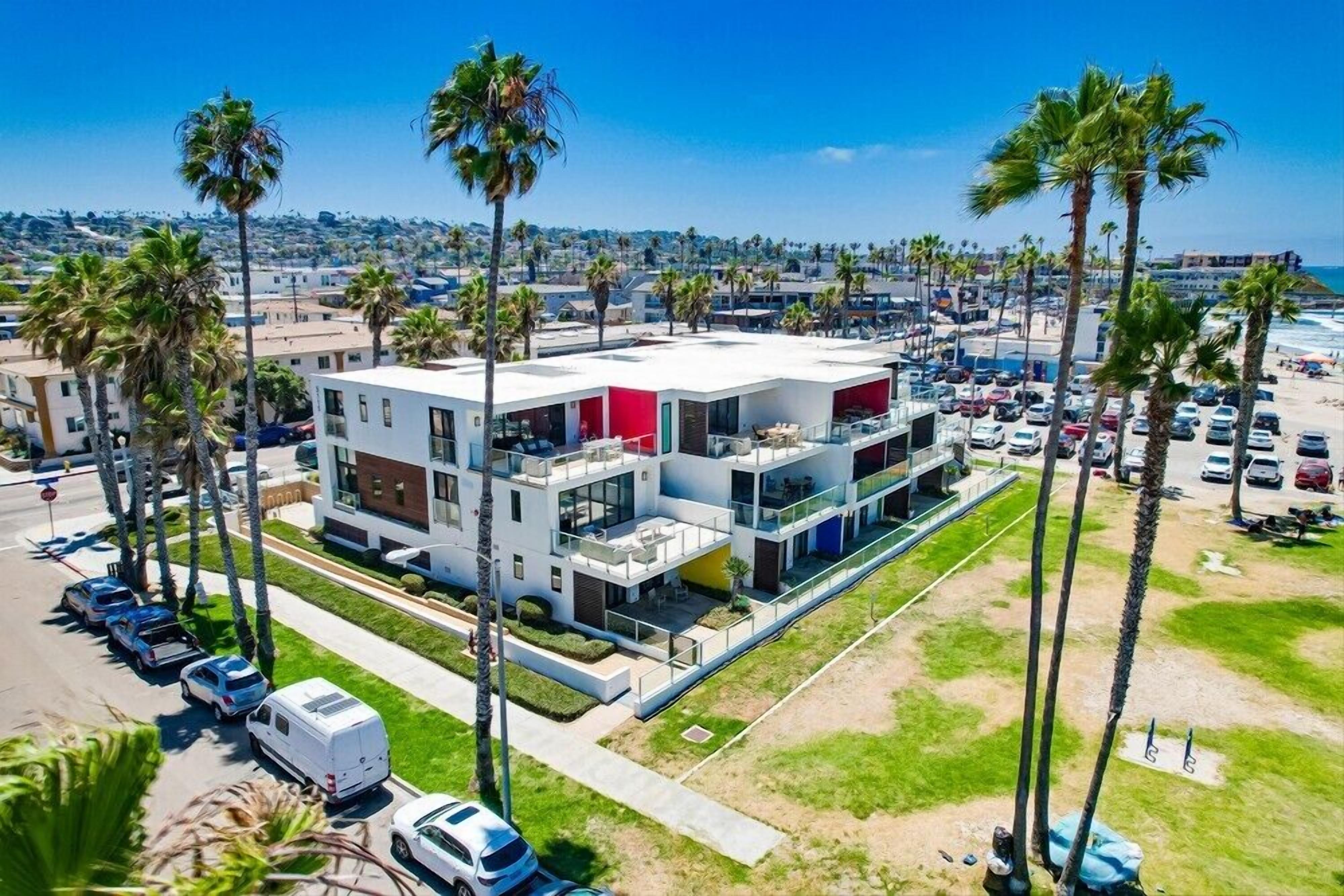 A multi-story hotel with a modern design, located near a beach with palm trees and a parking lot.