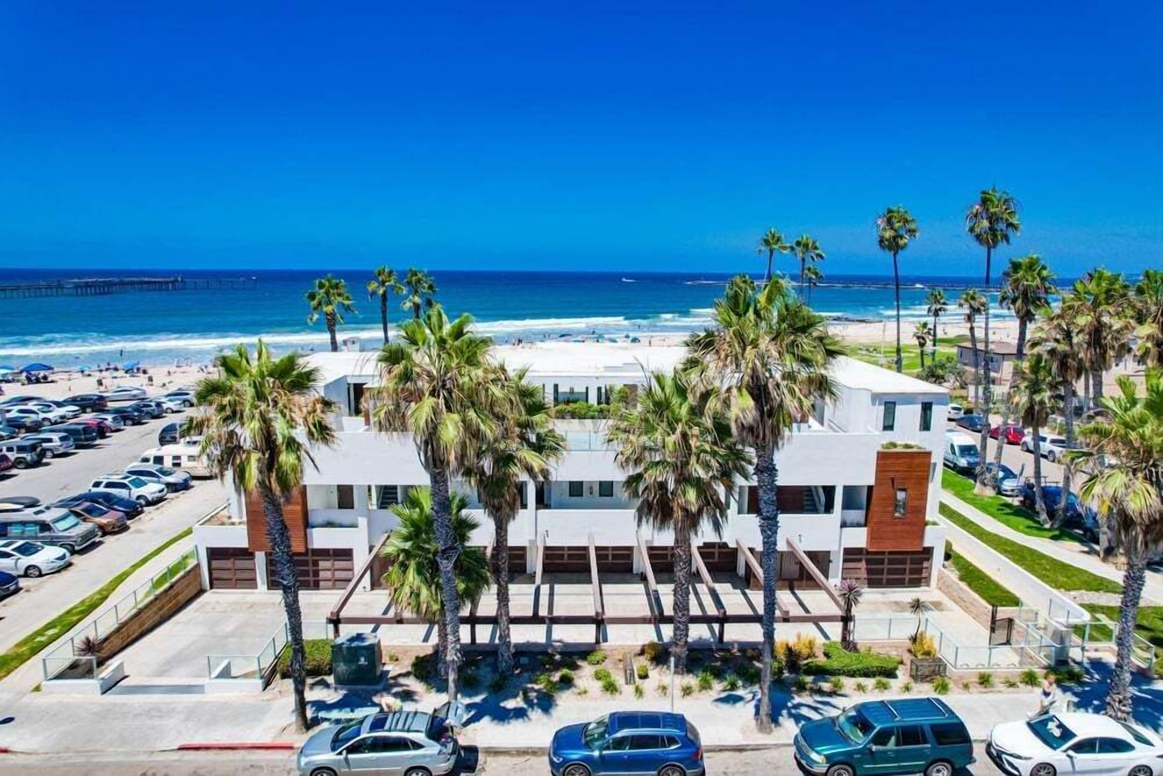 A modern hotel with a parking lot, palm trees, and a view of the beach and ocean.