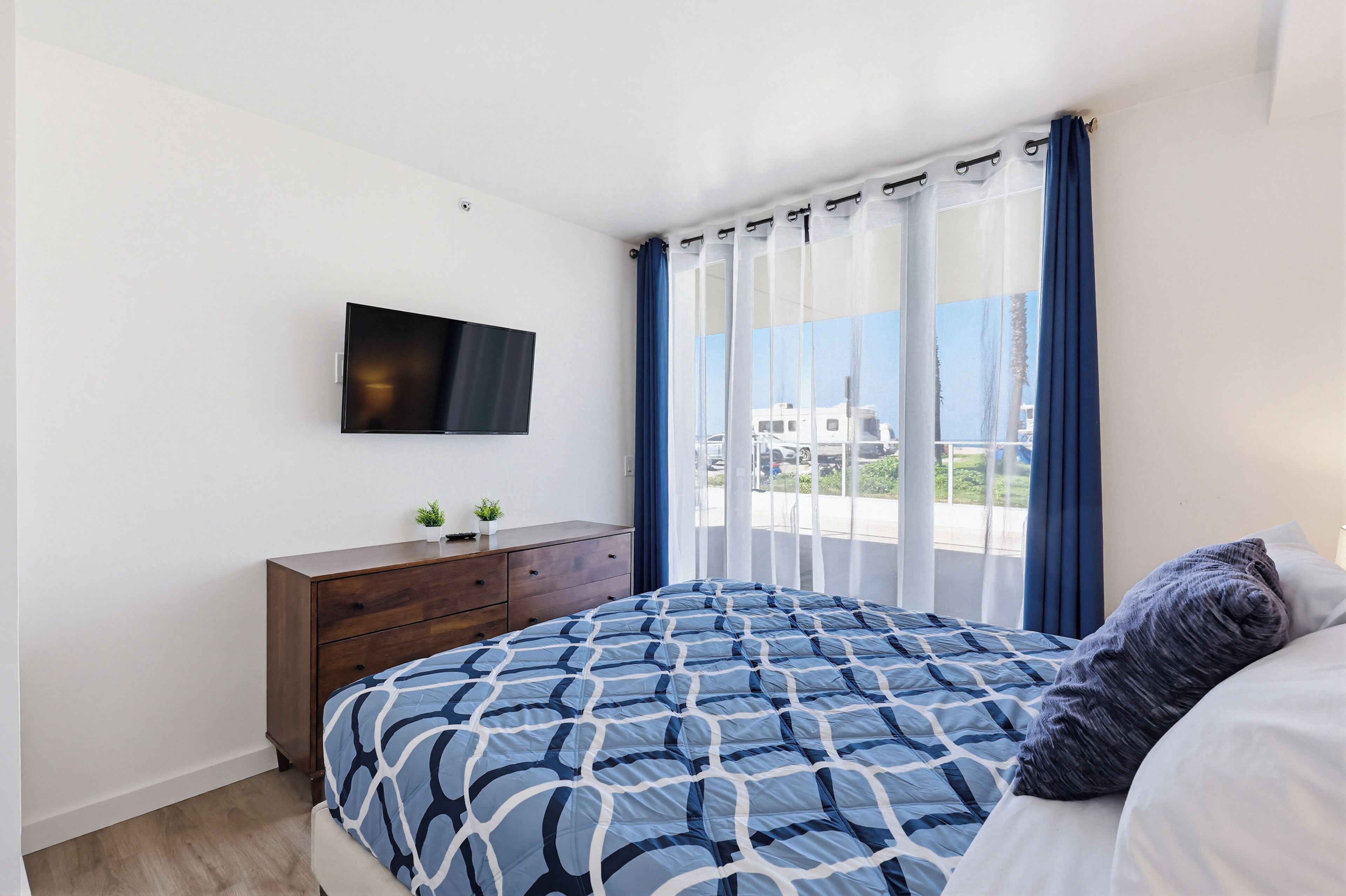 A bedroom with a bed, a TV mounted on the wall, a dresser, and a window with curtains.