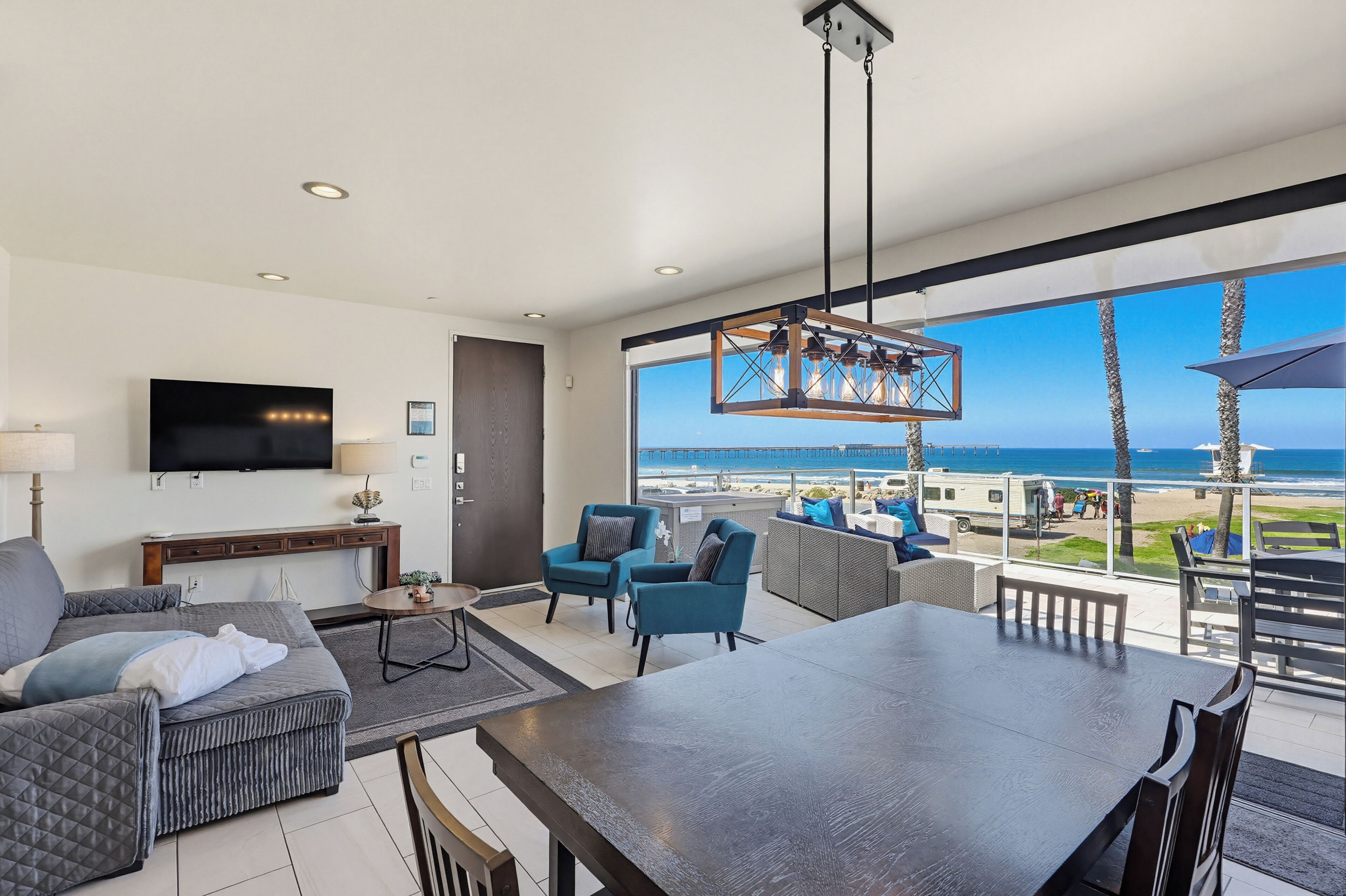 A modern living room with a large TV, comfortable seating, and a view of the beach.