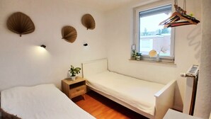 Una habitación pequeña con cama individual, una mesita de noche de madera, una planta en maceta y una ventana con vistas al exterior.