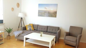 Una sala de estar con un sofá gris, una mesita de centro blanca, un sillón gris, una lámpara y una planta pequeña.