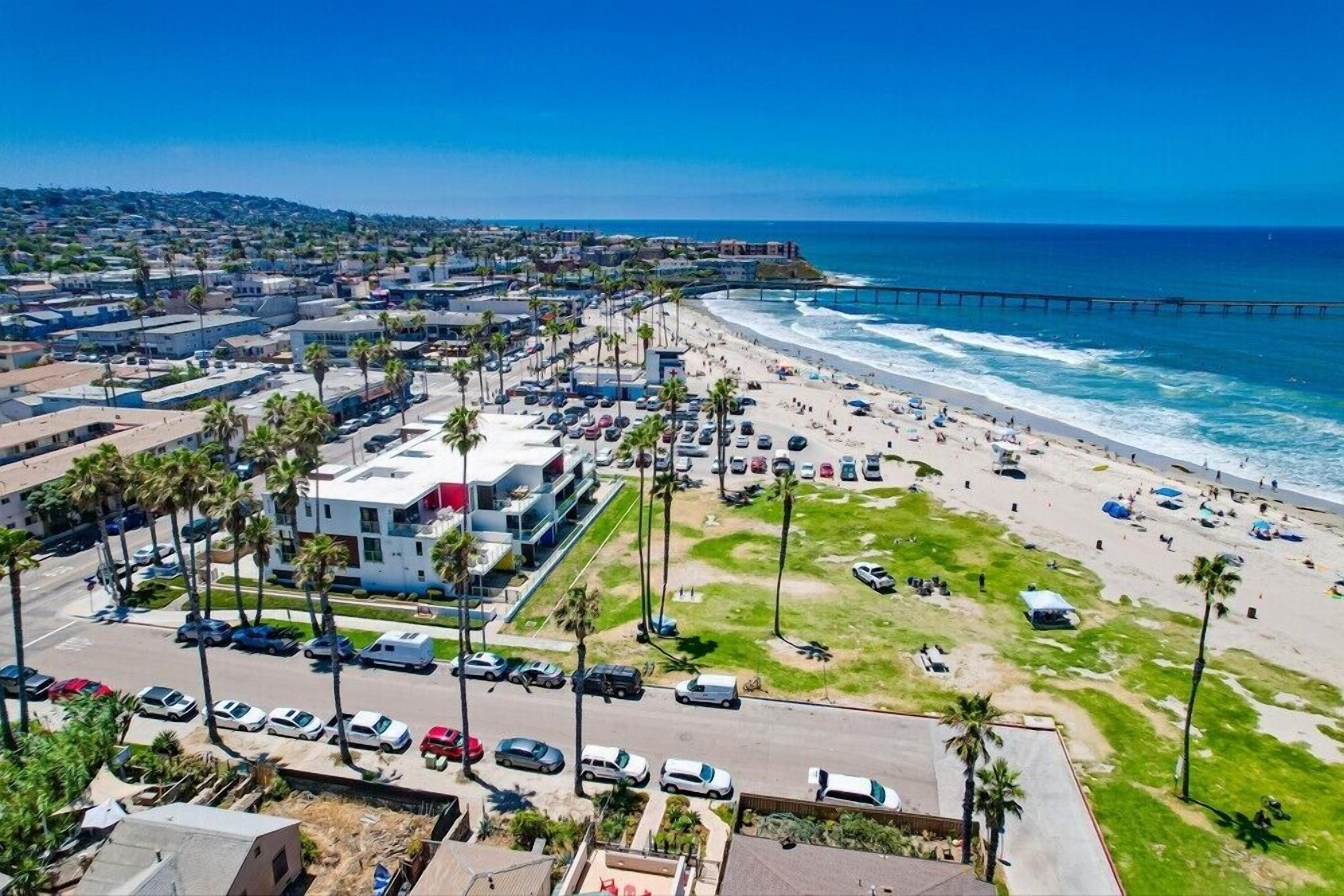 An aerial view of a coastal city with a beach, a pier, and a parking lot with cars and palm trees.
