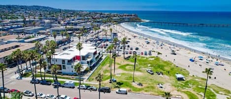 An aerial view of a coastal city with a beach, a pier, and a parking lot with cars and palm trees.