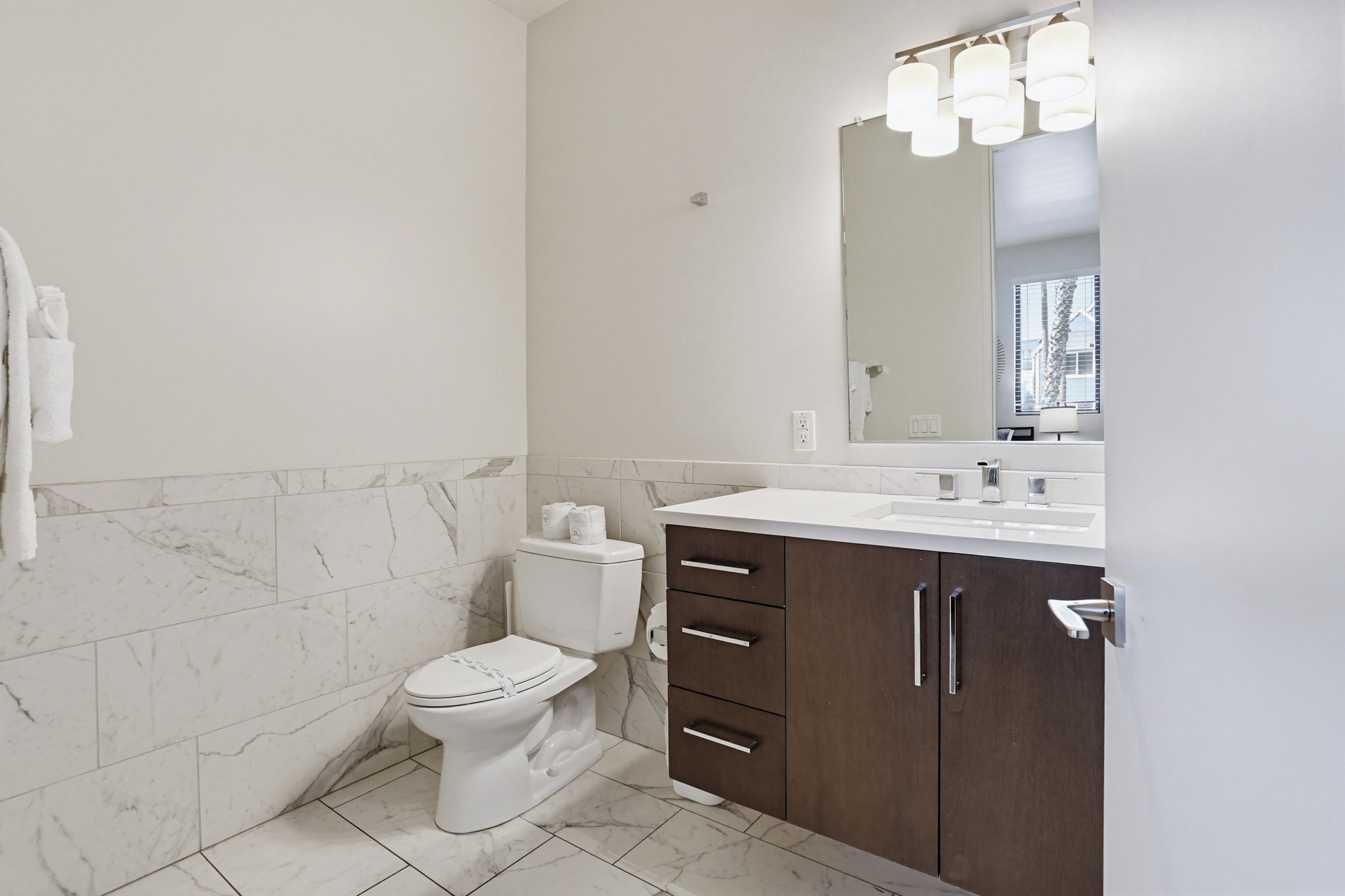 A bathroom with a toilet, a marble-tiled wall, a white sink, and a mirror with lights.