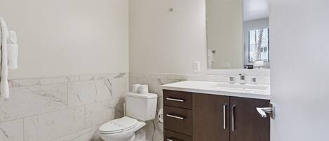 A bathroom with a toilet, a marble-tiled wall, a white sink, and a mirror with lights.