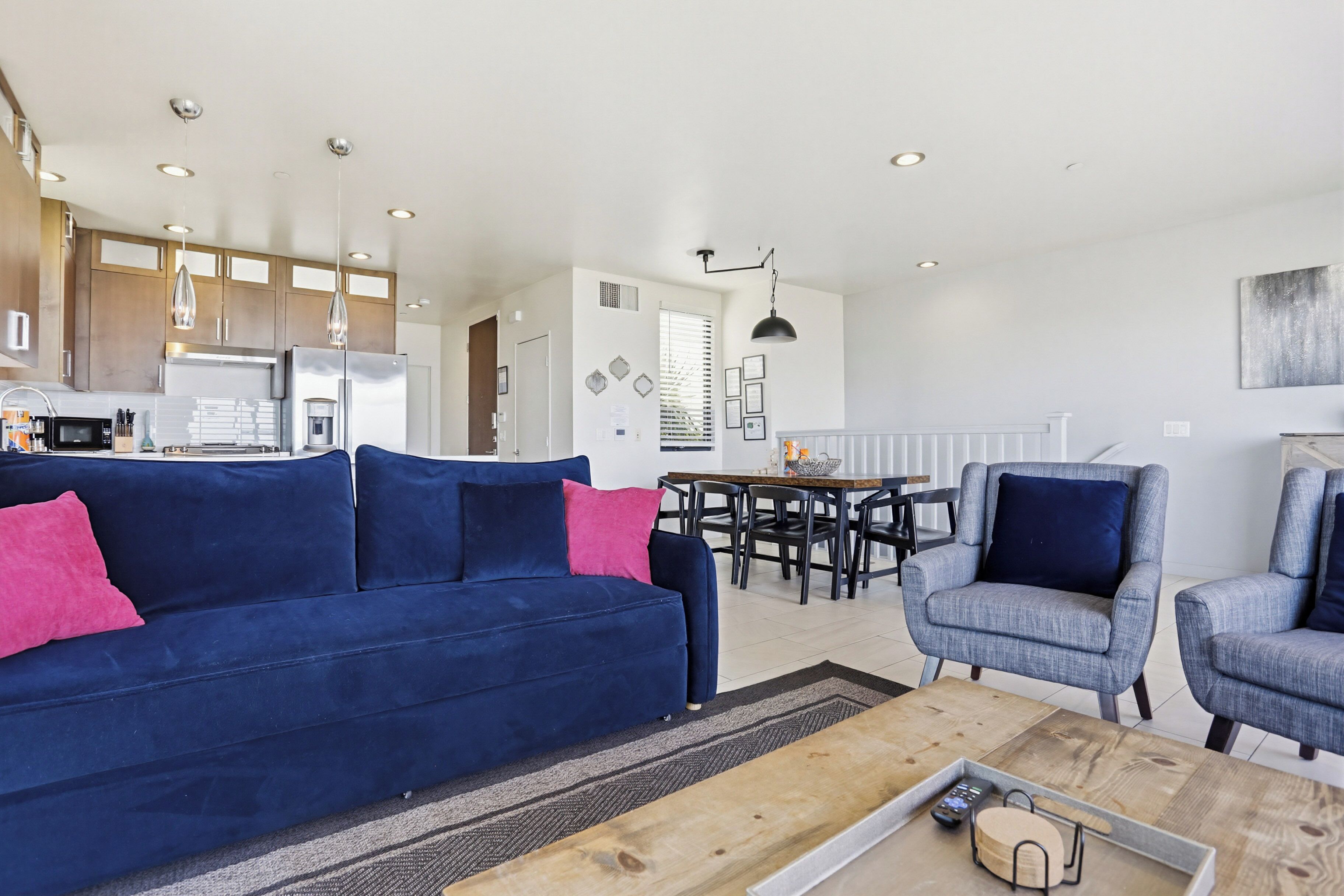 Een moderne woonkamer met een blauwe bank, grijze fauteuils, een houten salontafel en een eethoek met een tafel en stoelen.