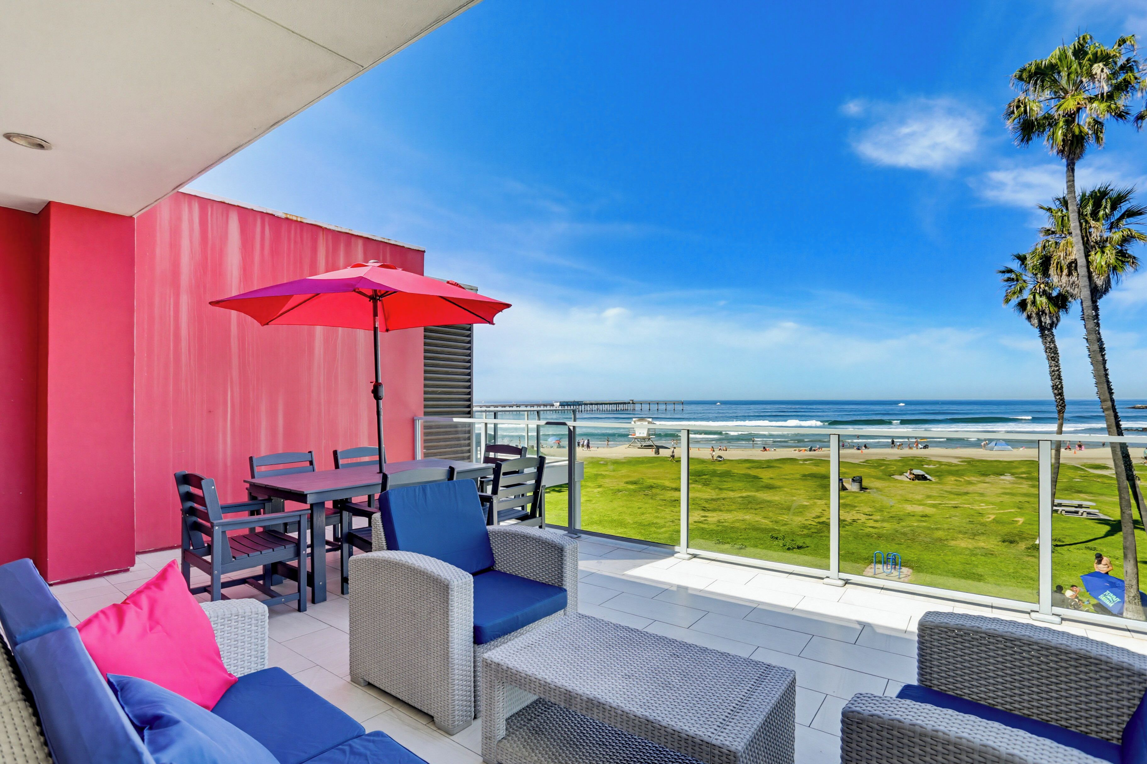 A balcony with wicker furniture, a red umbrella, and a view of the beach and ocean.