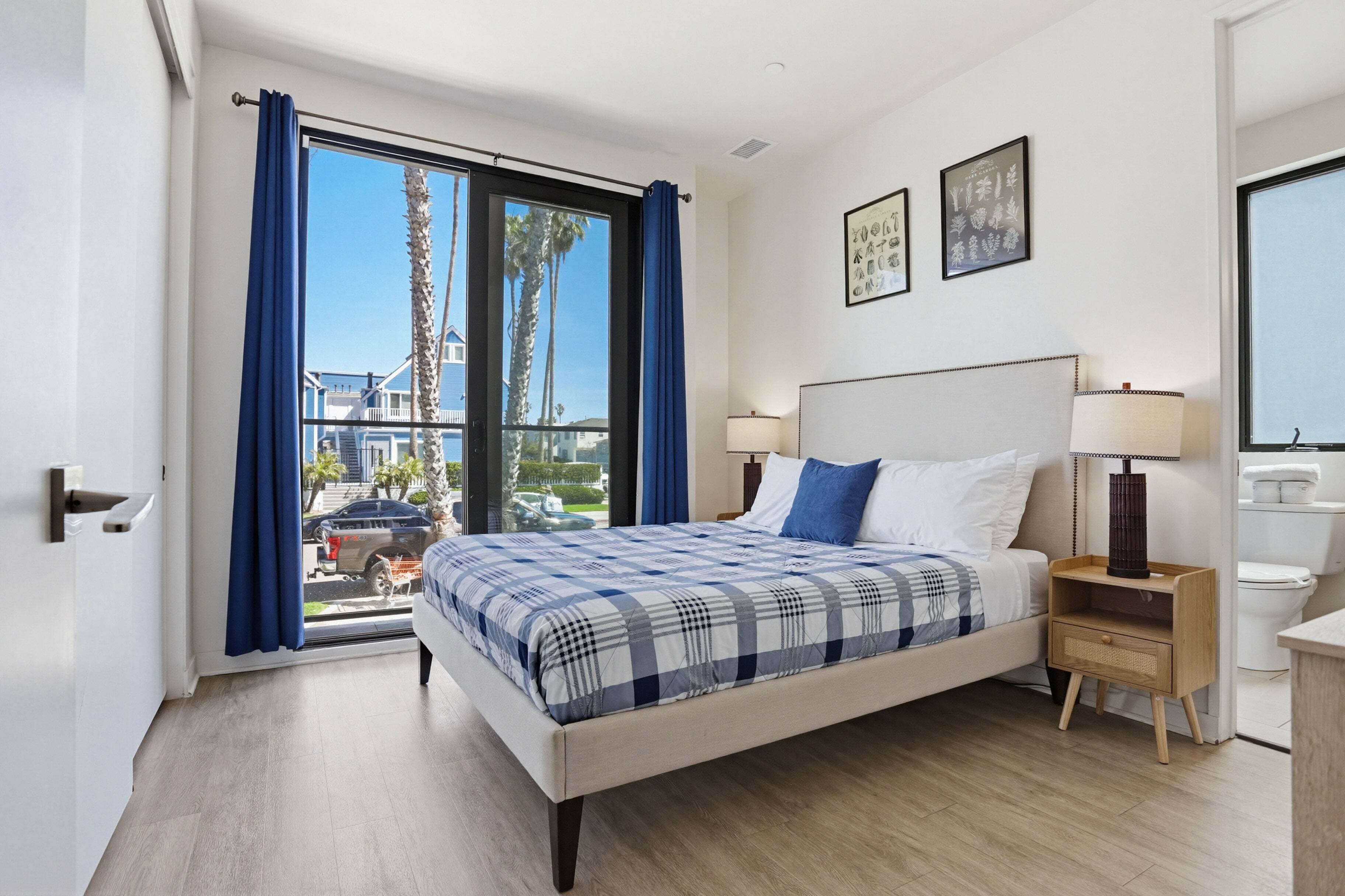 A modern hotel room with a large bed, a nightstand, a window with curtains, and a view of palm trees and buildings outside.