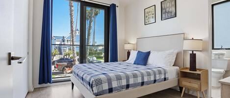 A modern hotel room with a large bed, a nightstand, a window with curtains, and a view of palm trees and buildings outside.