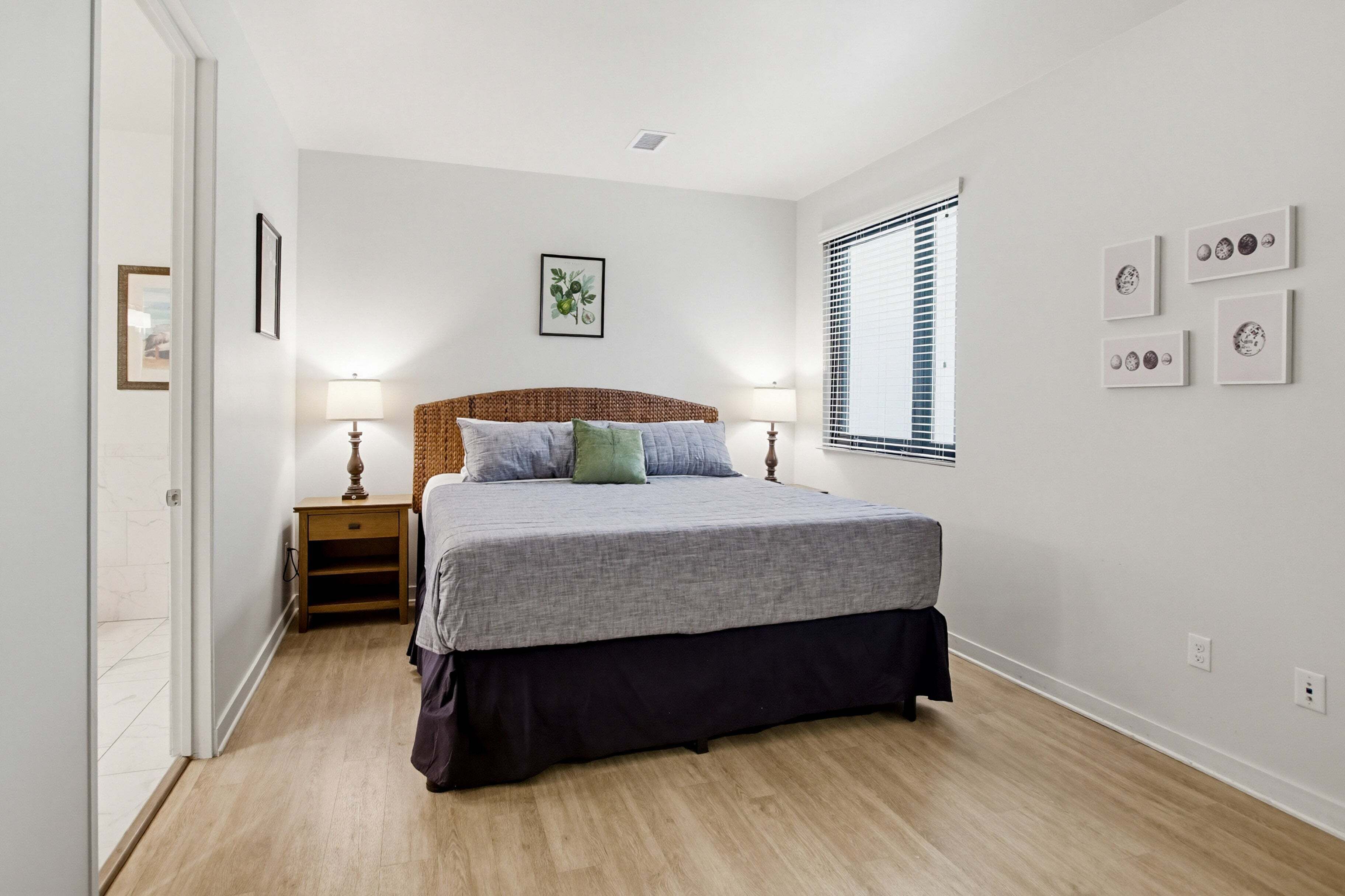 Une chambre à coucher comprenant un lit, des tables de chevet, une fenêtre avec des stores et des œuvres d’art accrochées au mur.