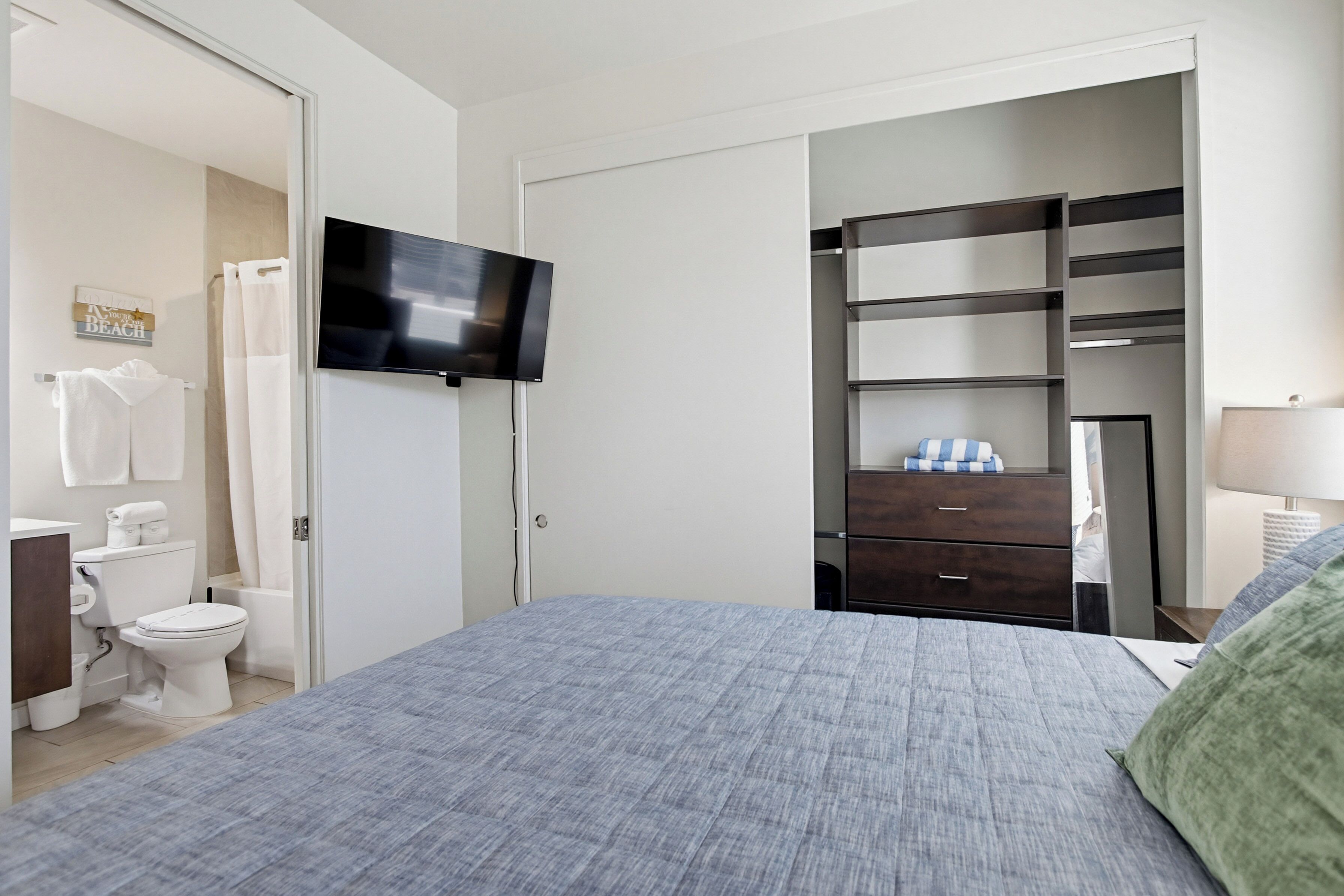 A bedroom with a bed, a TV, a wardrobe, a mirror, and a bathroom with a toilet and shower curtain.