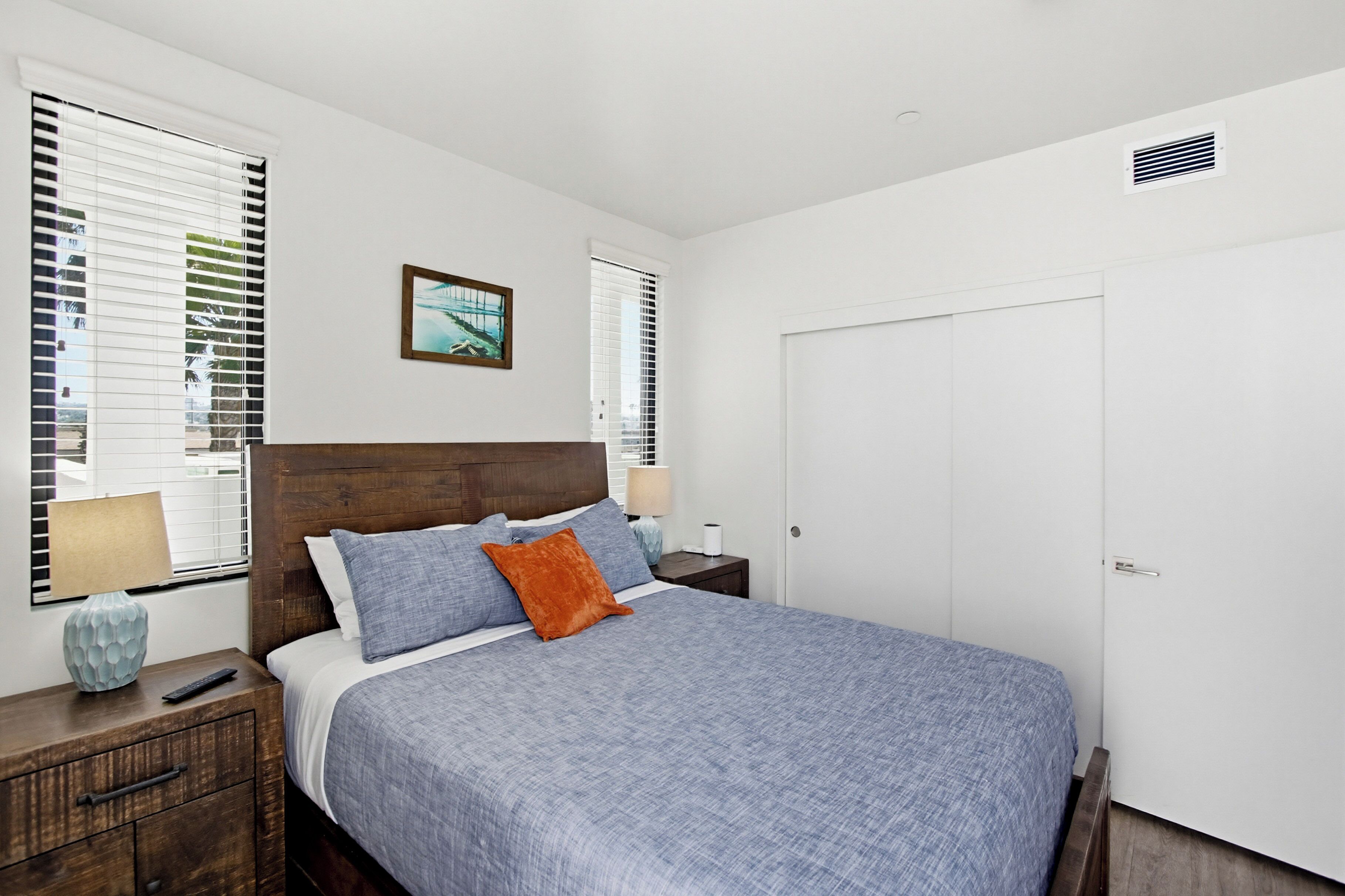 A bedroom with a bed, nightstands, a wooden headboard, a white door, and windows with blinds.