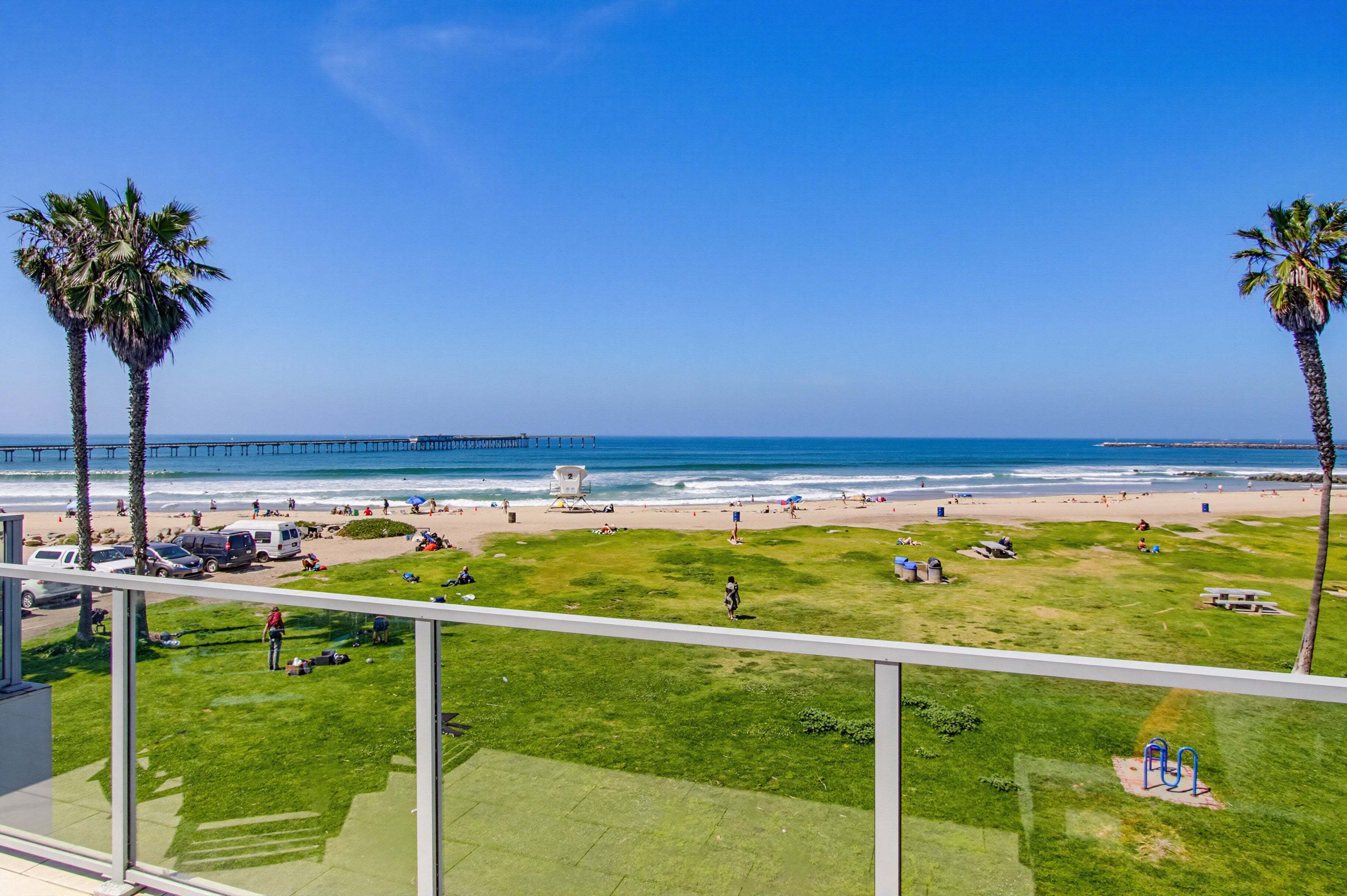 A beach view with palm trees, a pier, and a grassy area with people and vehicles.