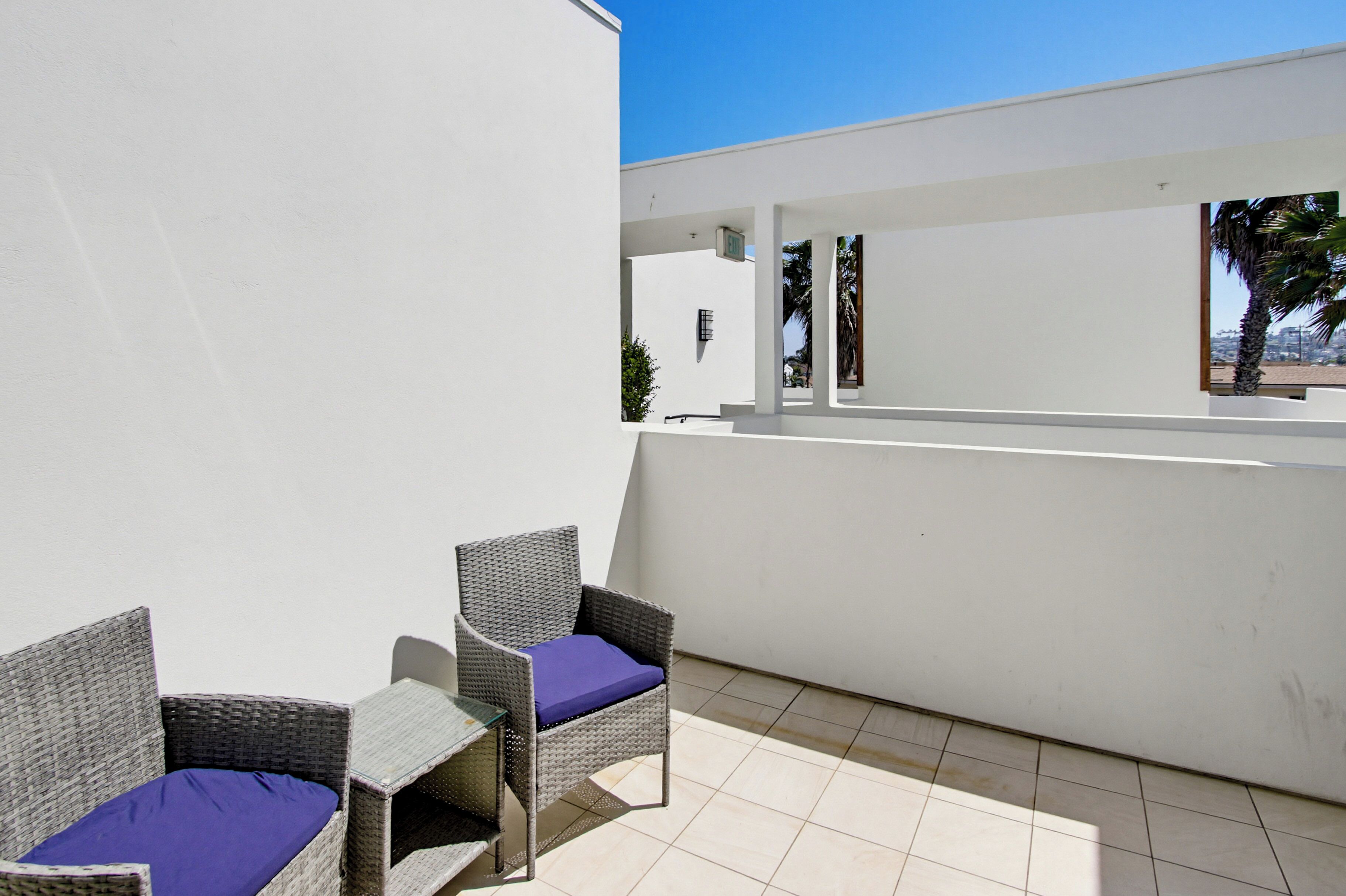 A balcony with wicker chairs and a small table, white walls, and a clear blue sky.