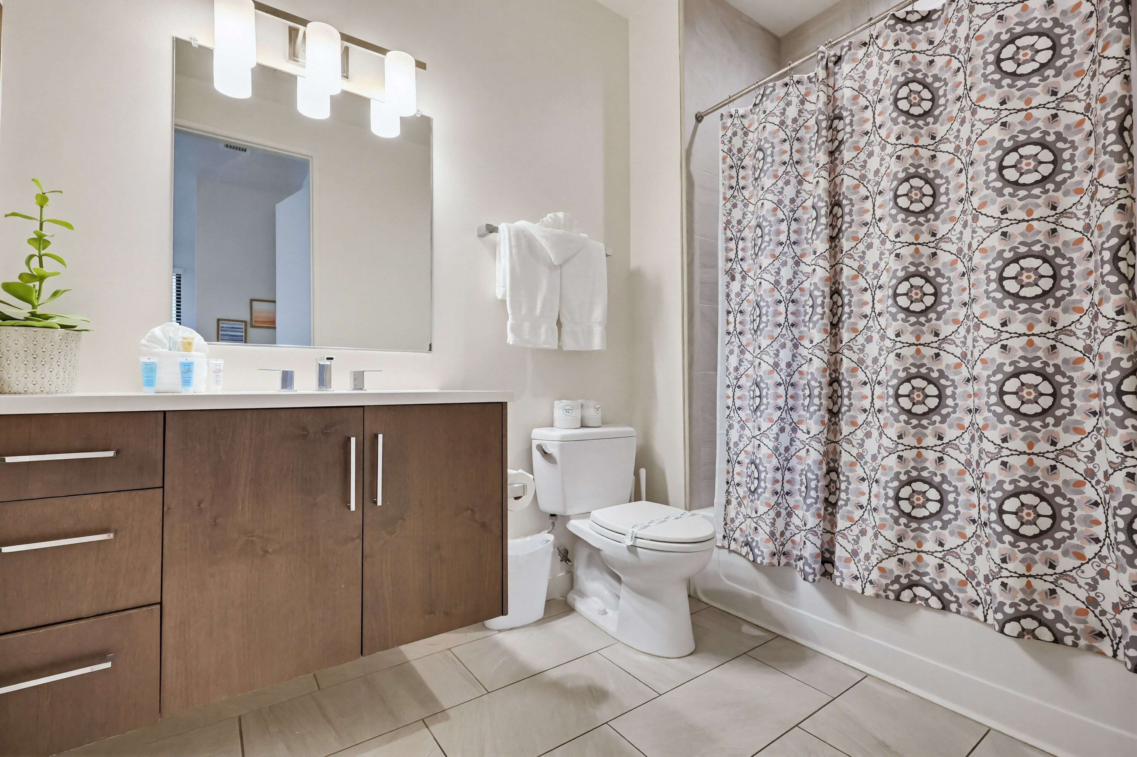 A bathroom with a wooden vanity, a white toilet, a shower curtain with a geometric pattern, and a mirror with lights above.