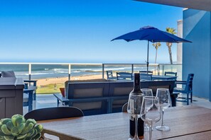 A table with a bottle and glasses, a blue umbrella, and a view of the beach.