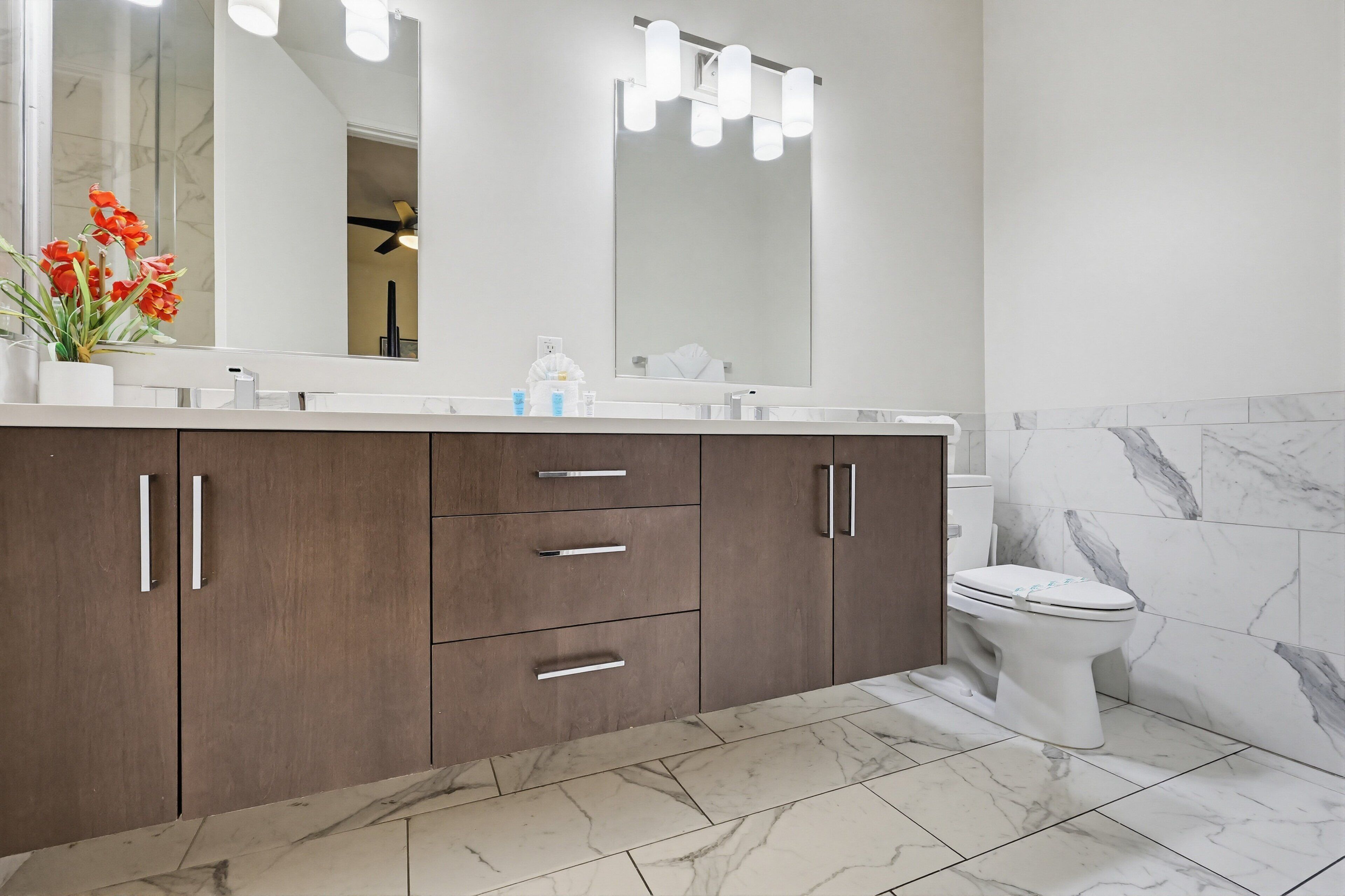 A bathroom with a marble floor, a white toilet, a wooden vanity with drawers, a large mirror with lights, and a vase with red flowers.
