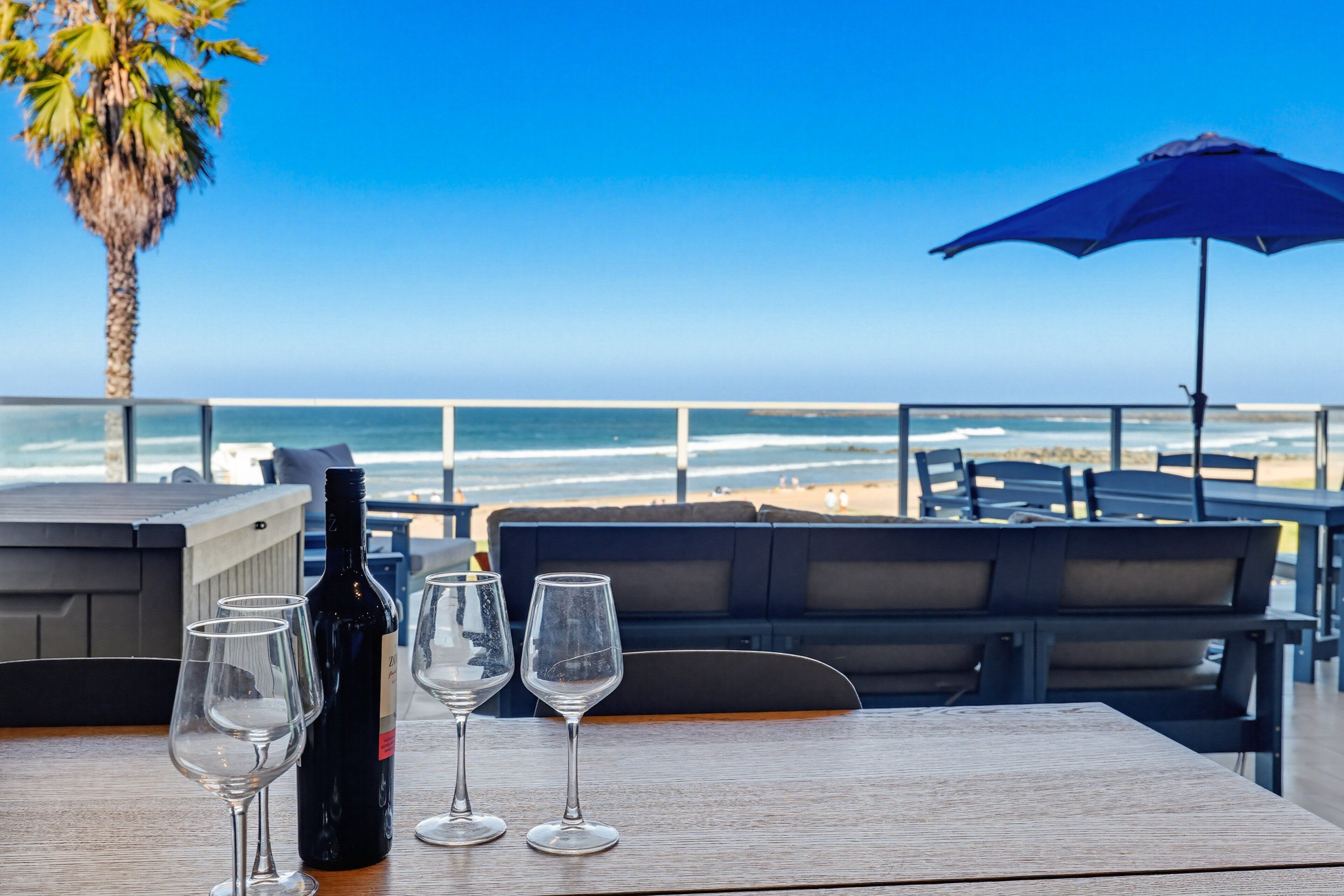 Ein Tisch mit einer Flasche und Gläsern, mit Blick auf einen Strand, der eine Palme und einen blauen Sonnenschirm zeigt.