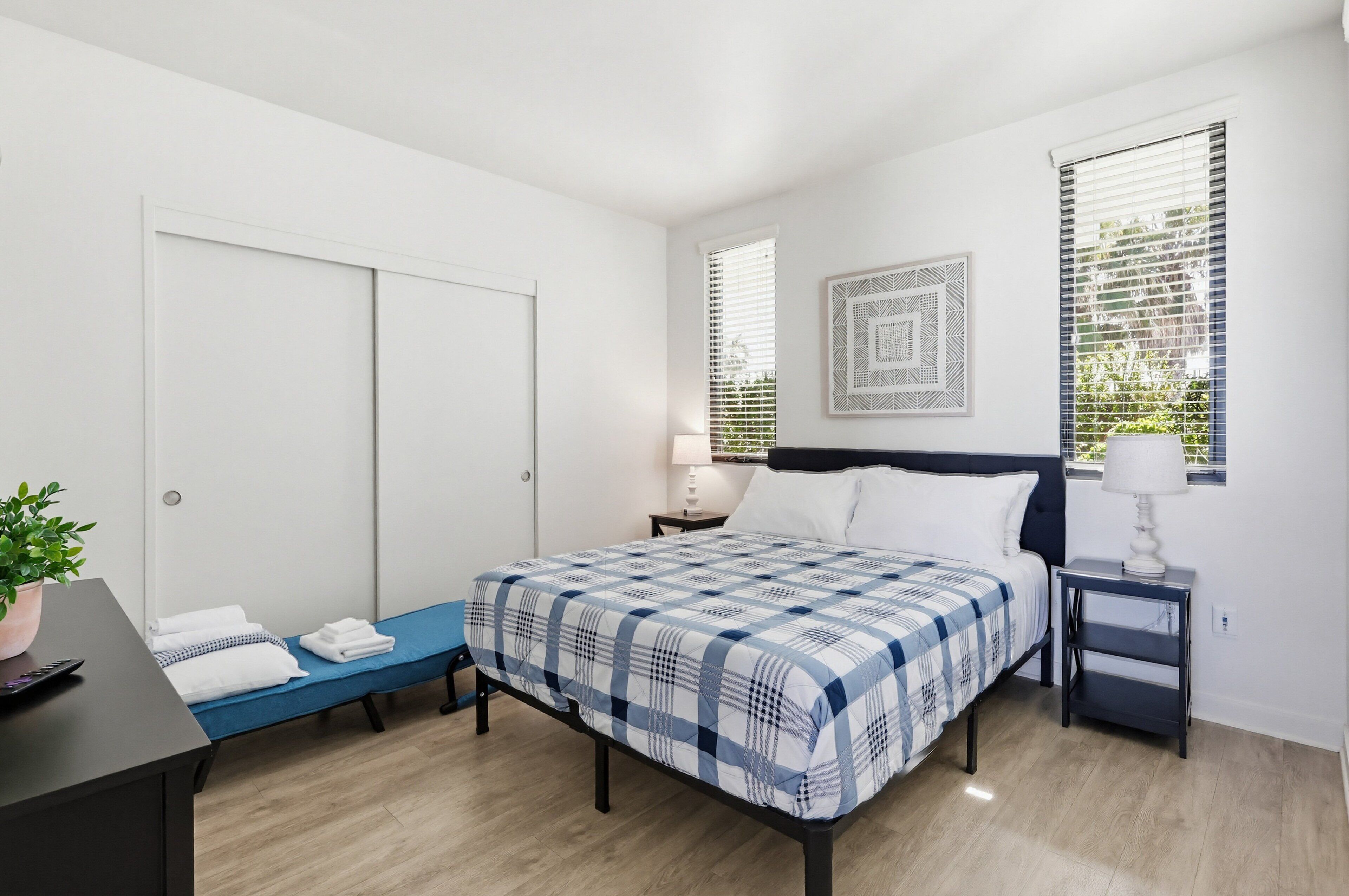 A bedroom with a bed, bedside tables, a desk, a chair, a potted plant, and a window with blinds.