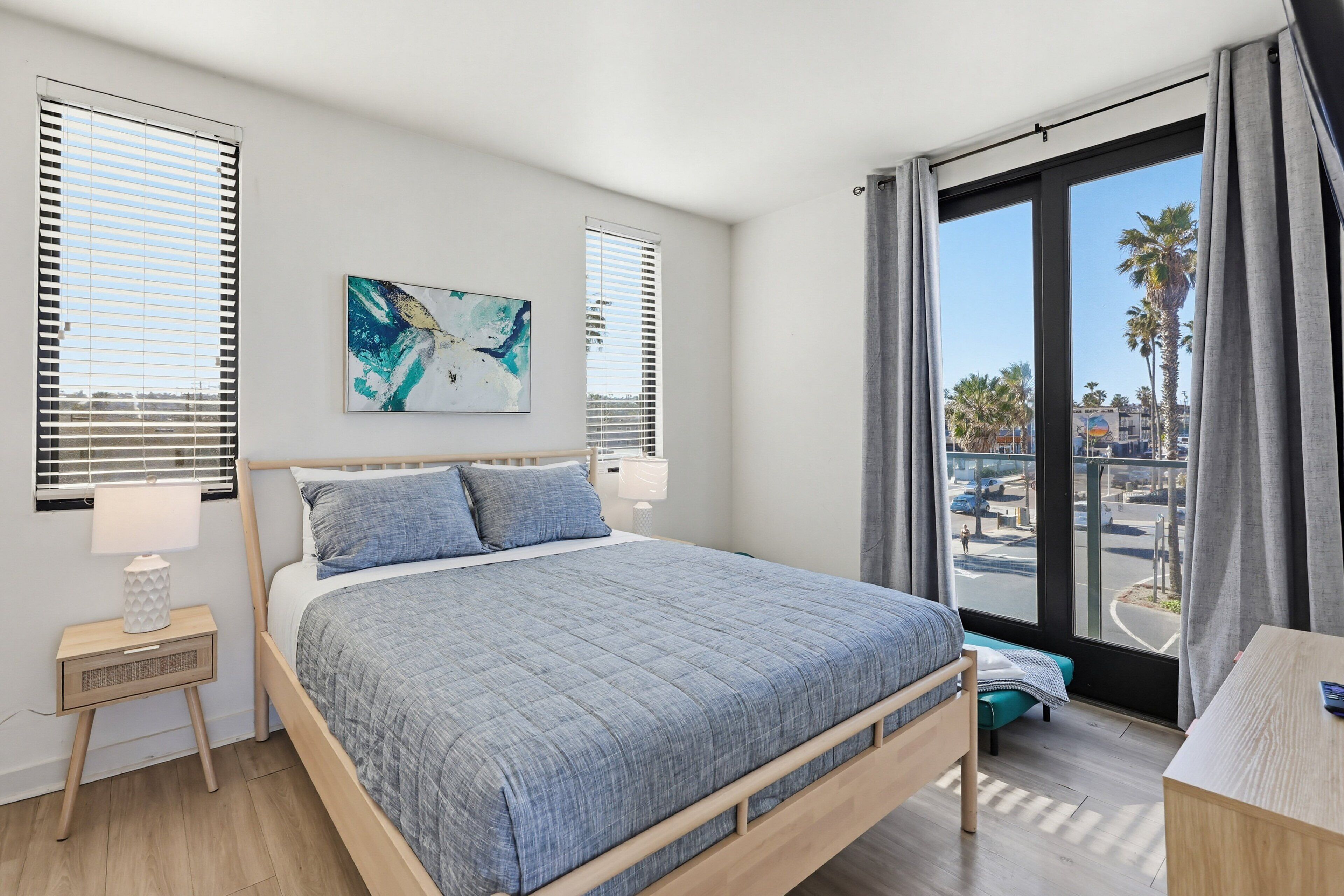 A bedroom with a bed, a nightstand, a painting, and a view of the street through large windows.
