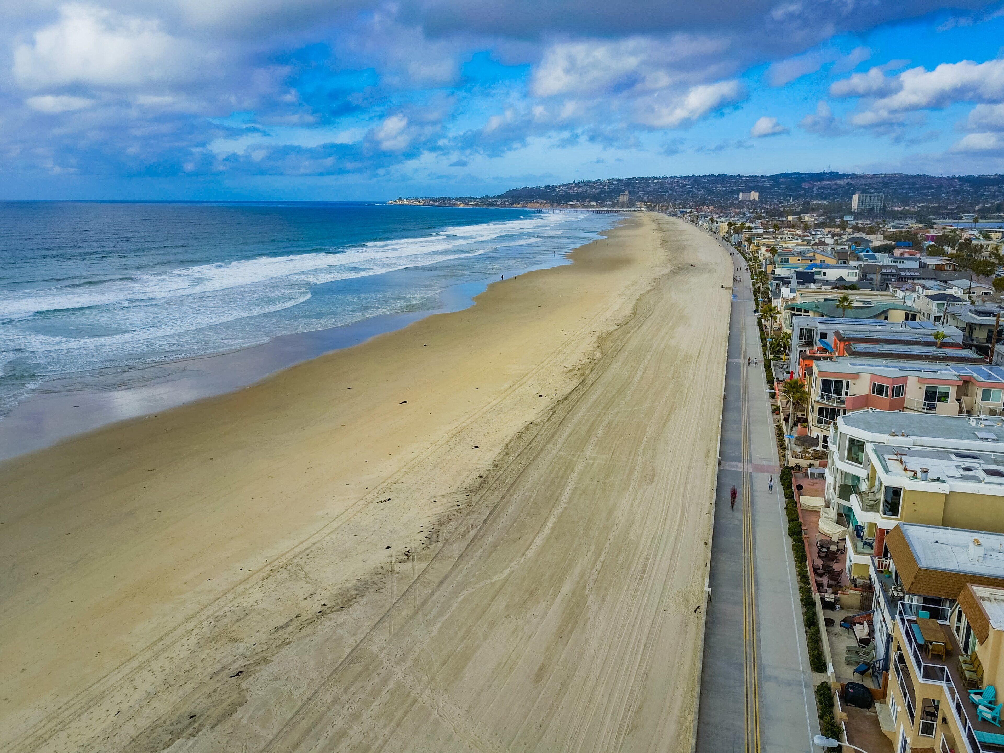 Vista costera con una extensa playa arenosa, un muelle que se adentra en el océano y una hilera de edificios a lo largo de la orilla.
