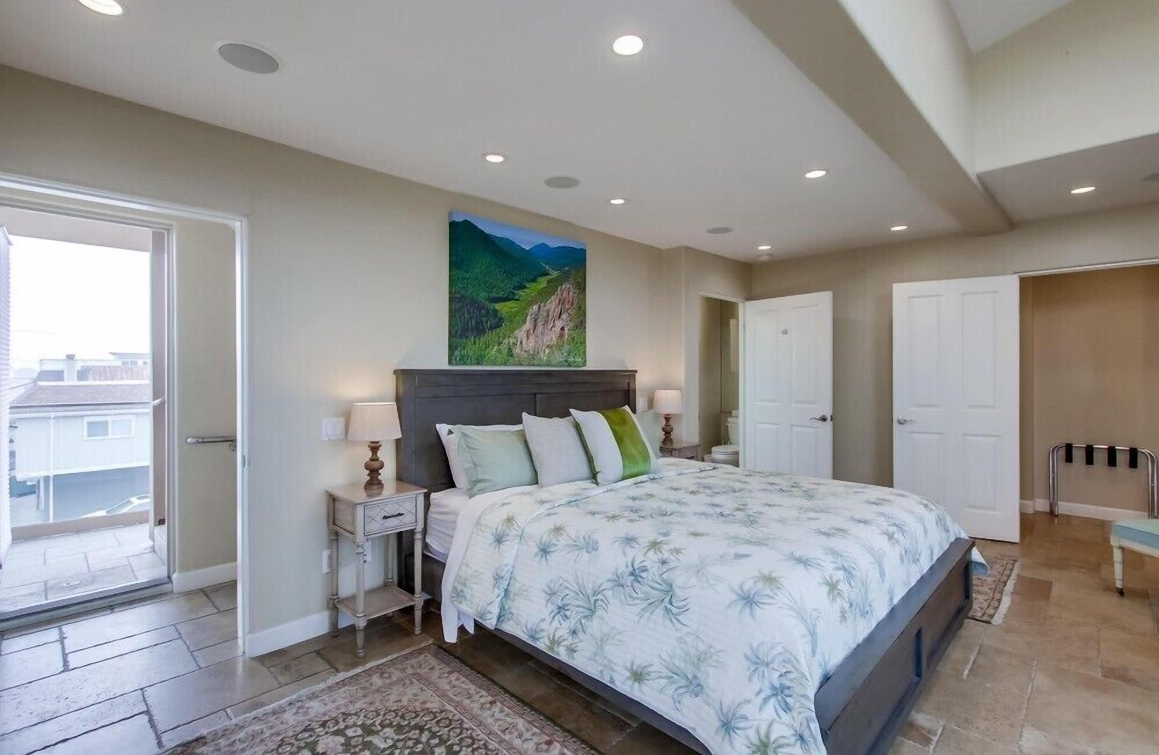 A bedroom with a large bed, a nightstand, a painting on the wall, and a door leading to another room.