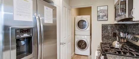 A compact kitchen with a stainless steel refrigerator, a built-in water dispenser, a double-door washer and dryer, a gas stove, and granite countertops.