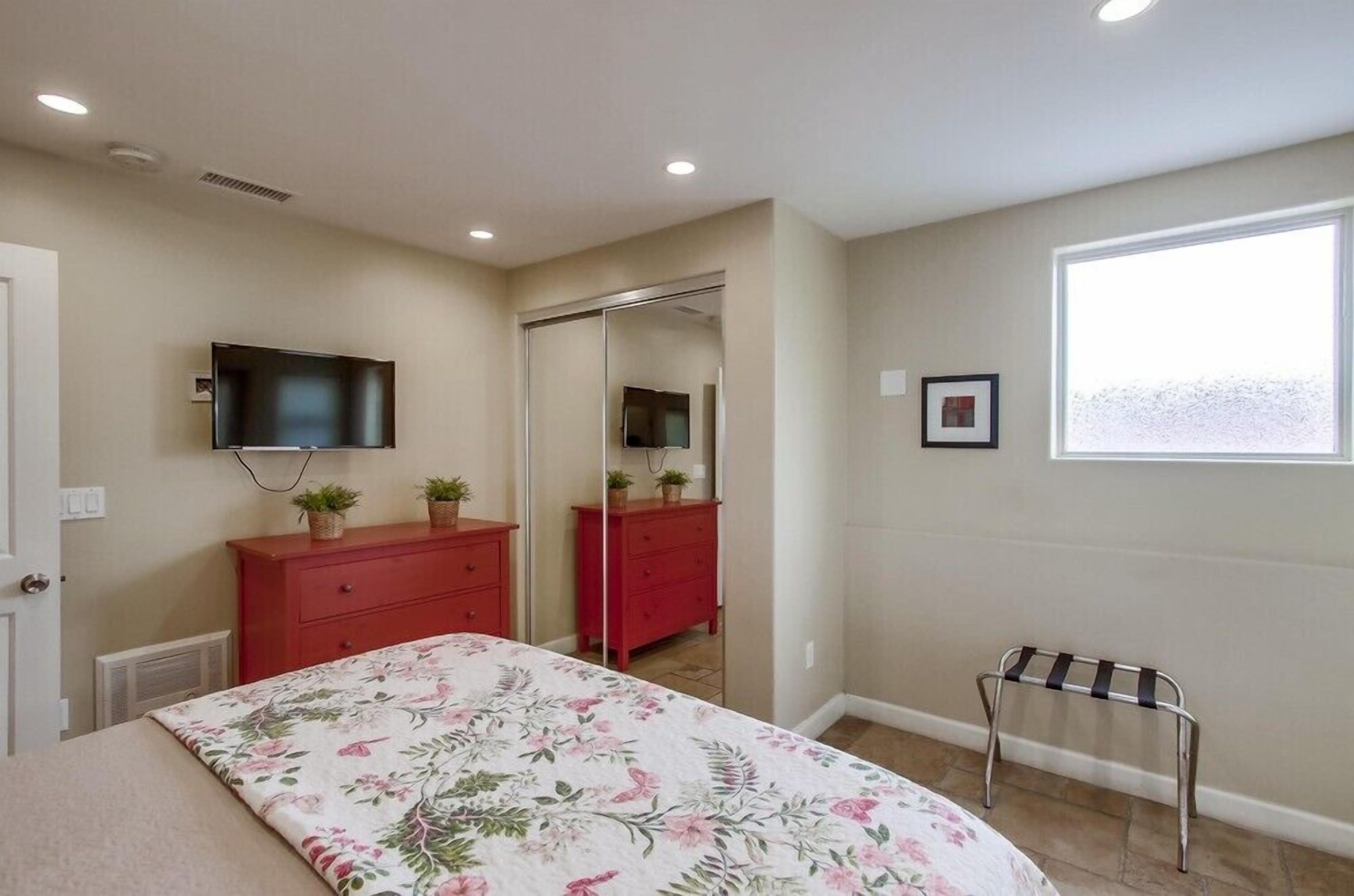 A bedroom with a bed, a red dresser, a TV mounted on the wall, a window, and a mirror.