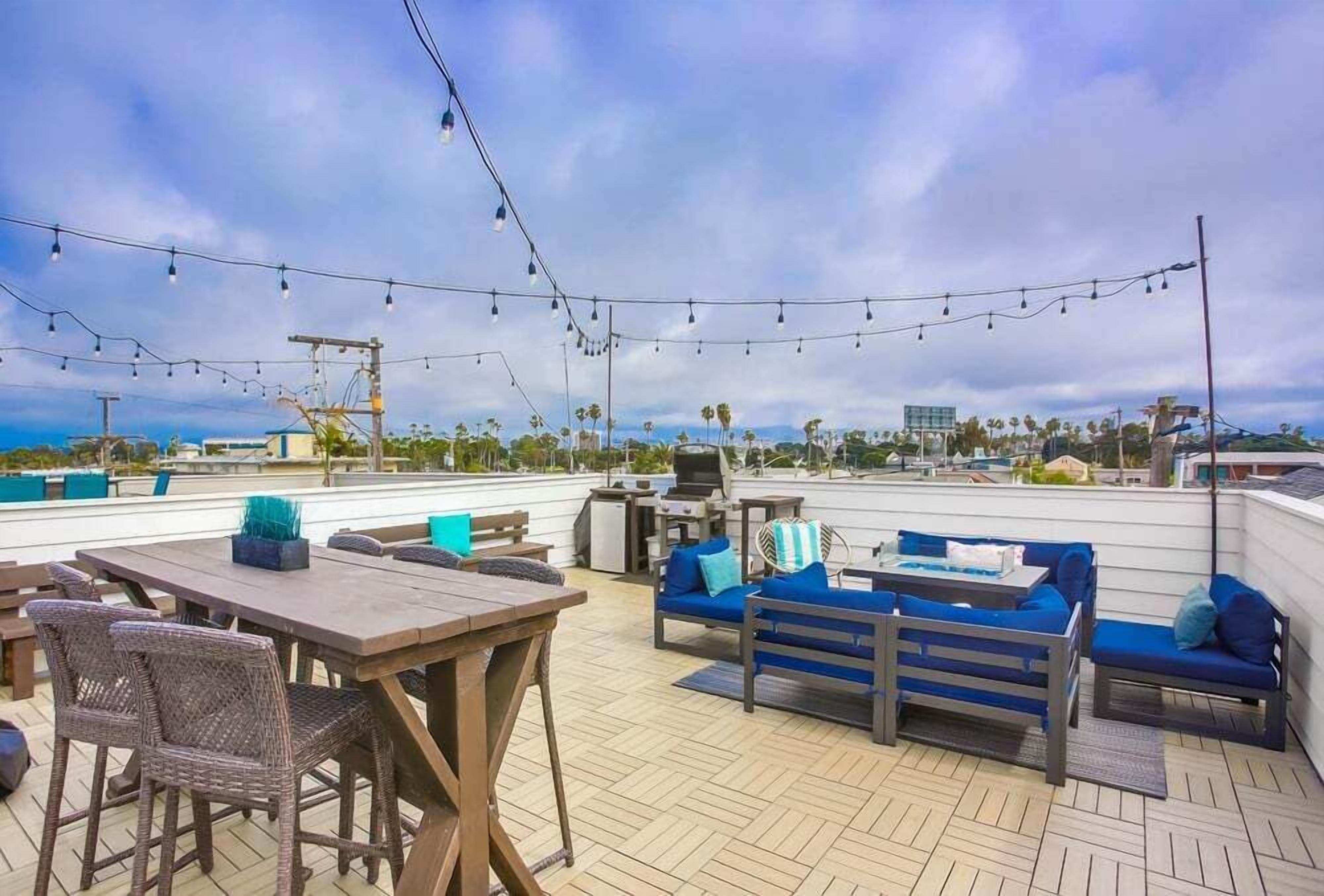 A rooftop patio with a wooden table, wicker chairs, blue sofas, and string lights.