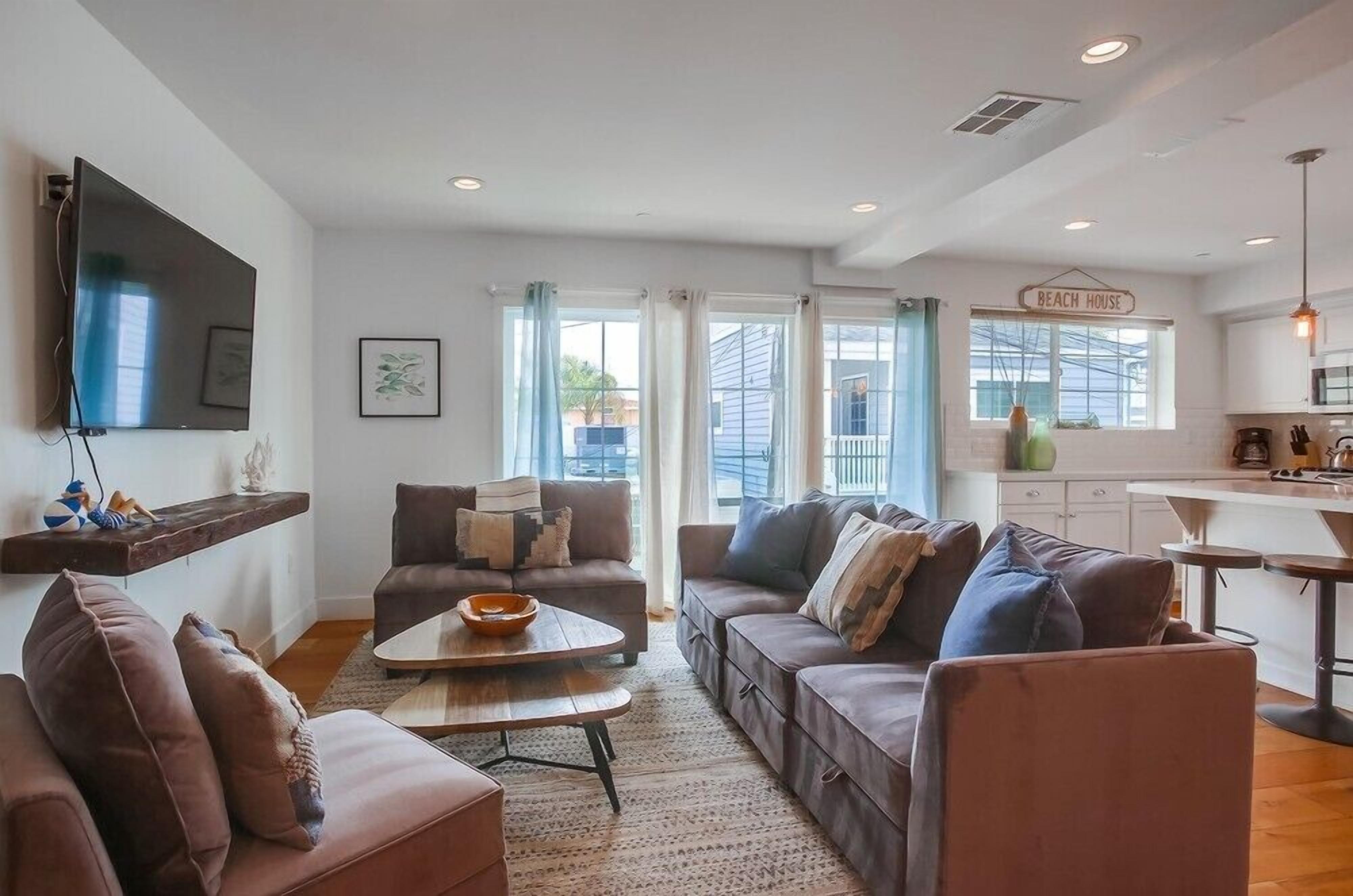 A modern living room with a flat-screen TV, two sofas, a coffee table, and a kitchen area with a sign reading "BEACH HOUSE".